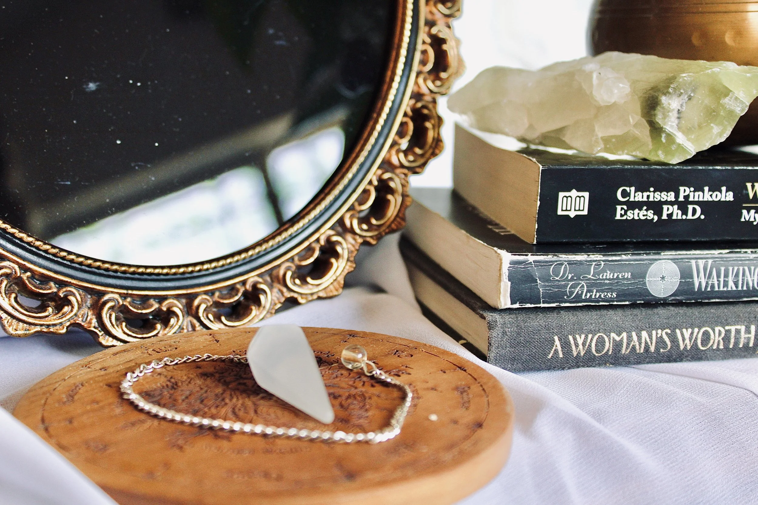 Close-up of a decorative mirror with gold and black ornate frame, placed on a surface with a jewelry chain in front of it. Behind the mirror, there are stacked books with visible titles, including one by Clarissa Pinkola Estés.