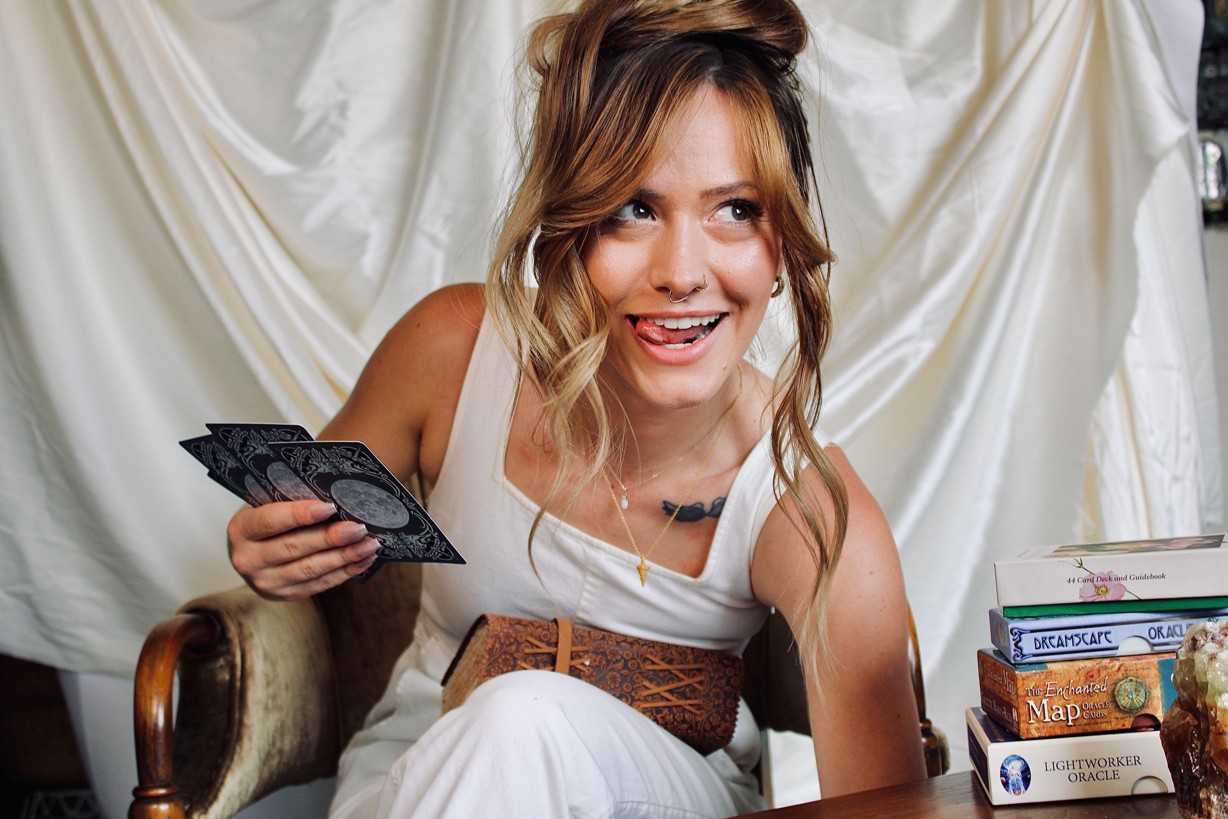 A young woman with brown hair, some dyed blonde, and a septum piercing, holding tarot cards in her right hand, smiling playfully with her tongue slightly out. She is sitting indoors, surrounded by books and crystals, with a white curtain in the background.