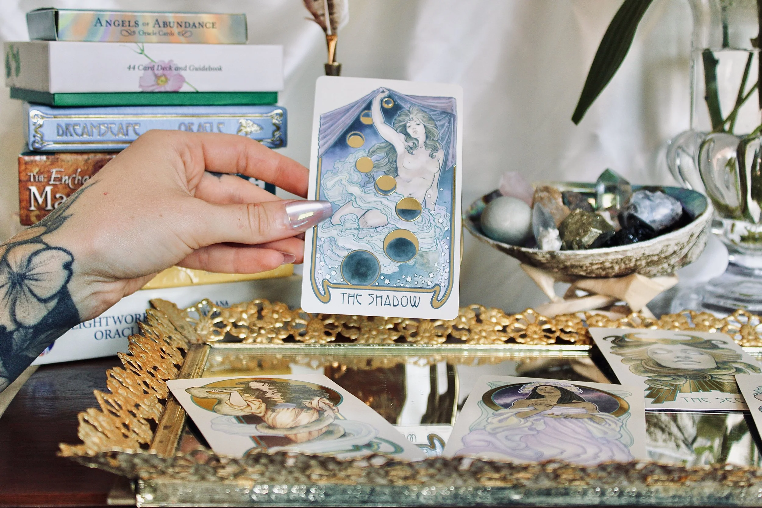 A hand holding a tarot card titled "The Shadow," featuring an illustrated woman with long flowing hair standing in water, with a curtain behind her and celestial motifs.