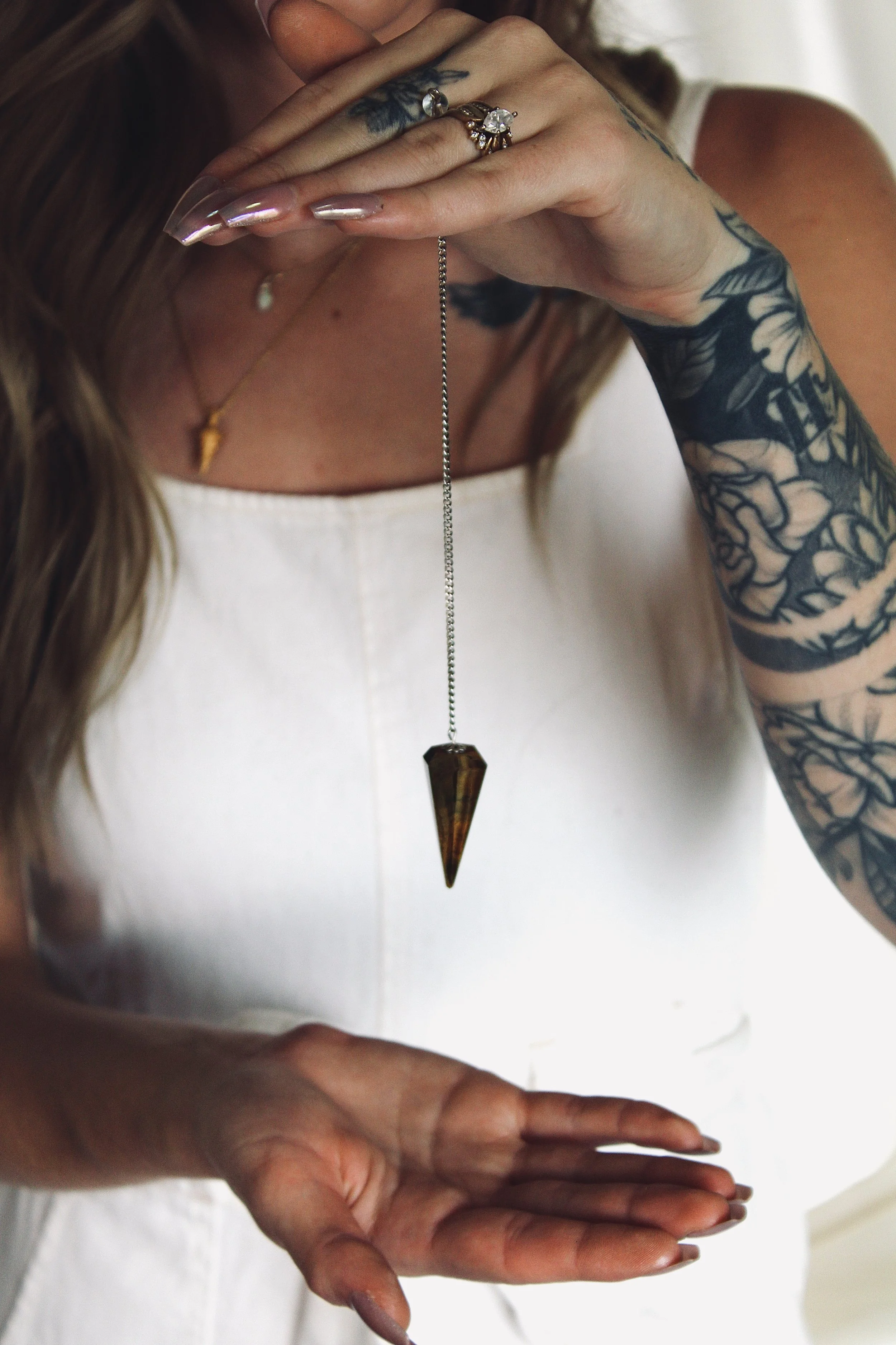 Close-up of a person holding their hands together in a prayer position, wearing rings and a necklace, and with a tattooed arm, against a blurred background.