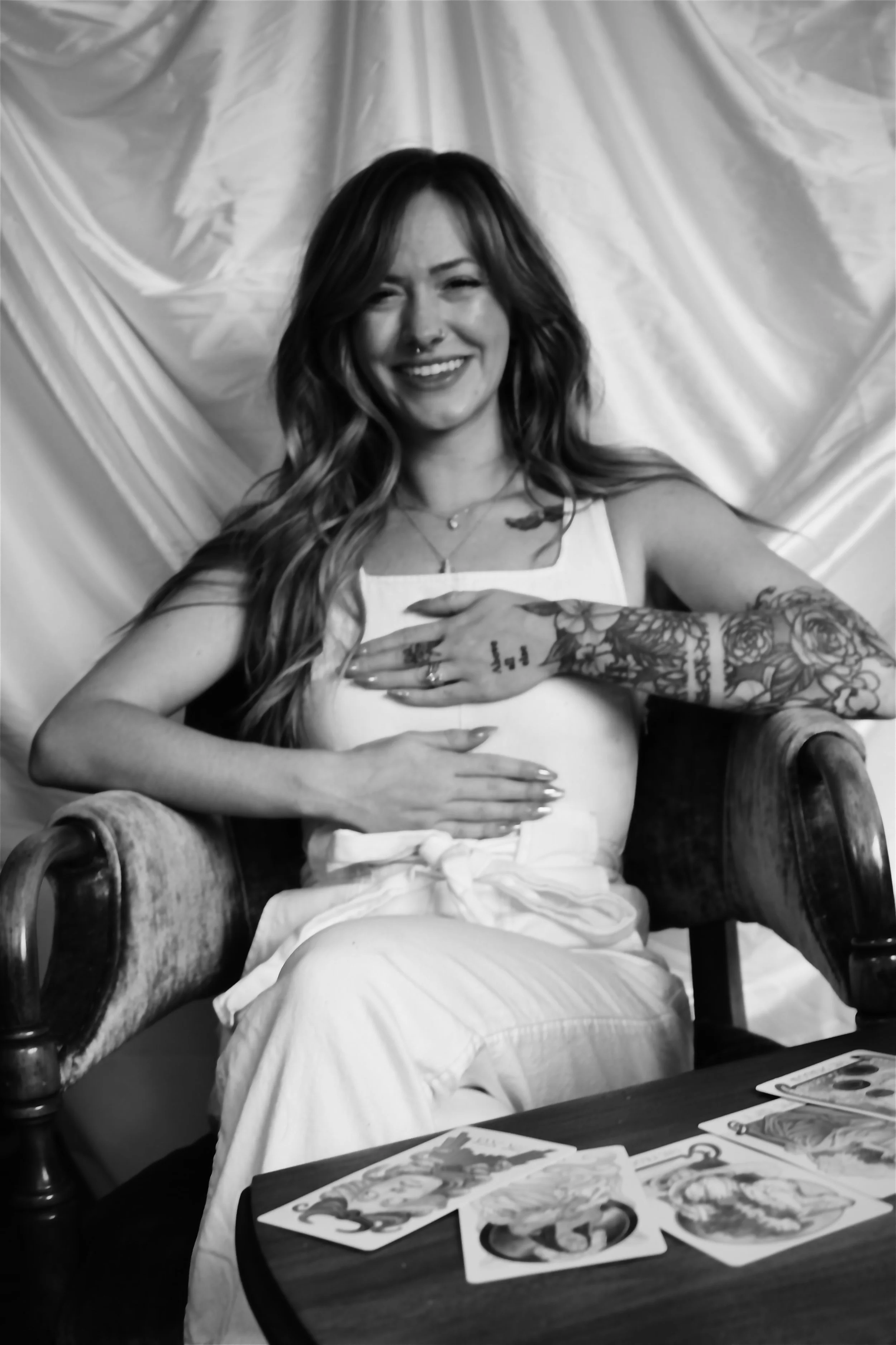 A smiling woman with tattoos on her right arm, sitting on a vintage wooden chair with ornate armrests, holding her stomach. She has long wavy hair and is dressed in a white sleeveless top. In front of her on a table is a spread of tarot cards, and the background features a crinkled fabric curtain.