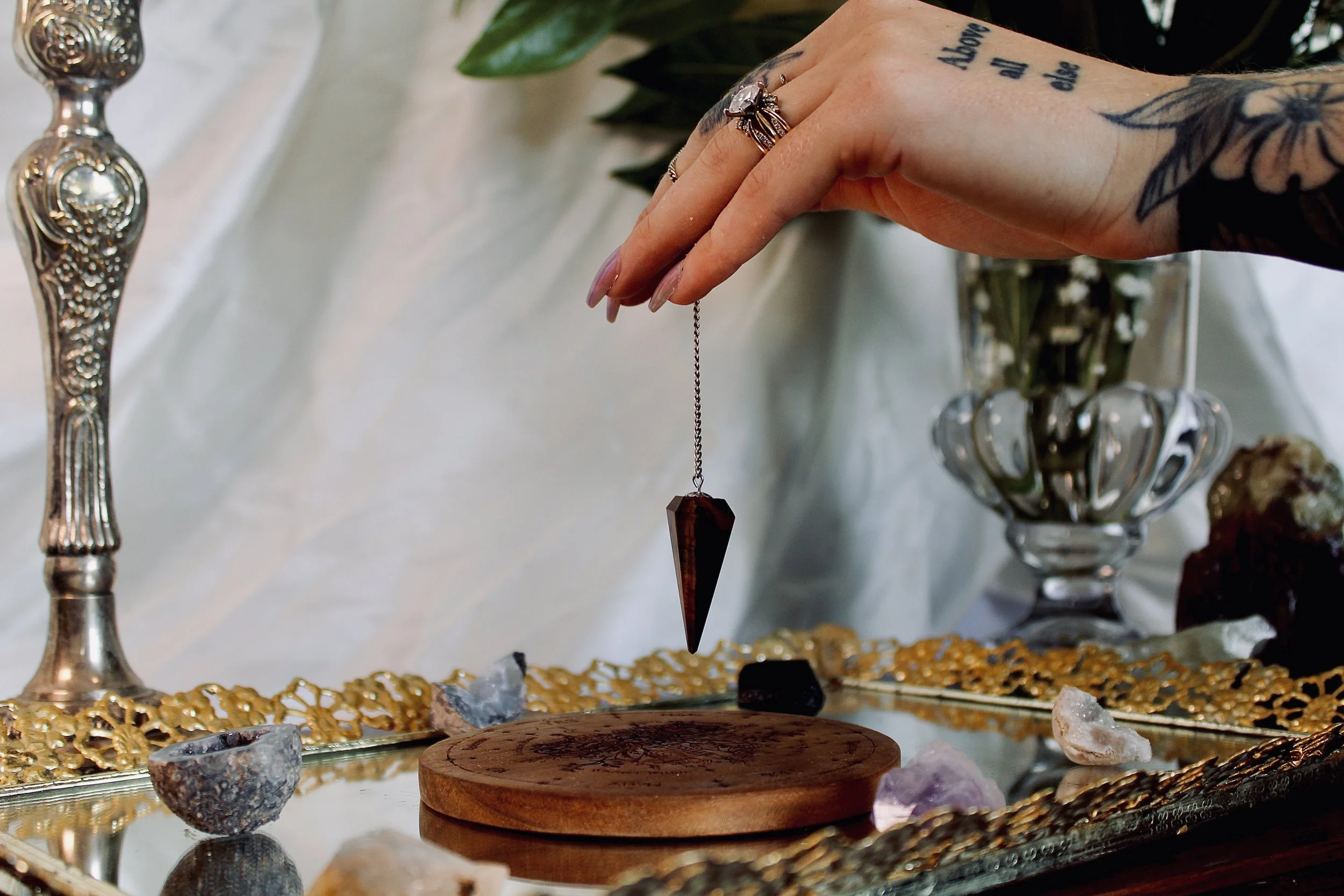 A person's hand with rings and tattoos reaching over a table with crystals, stones, and jewelry. The table has decorative items and a gold-colored ornate tray.