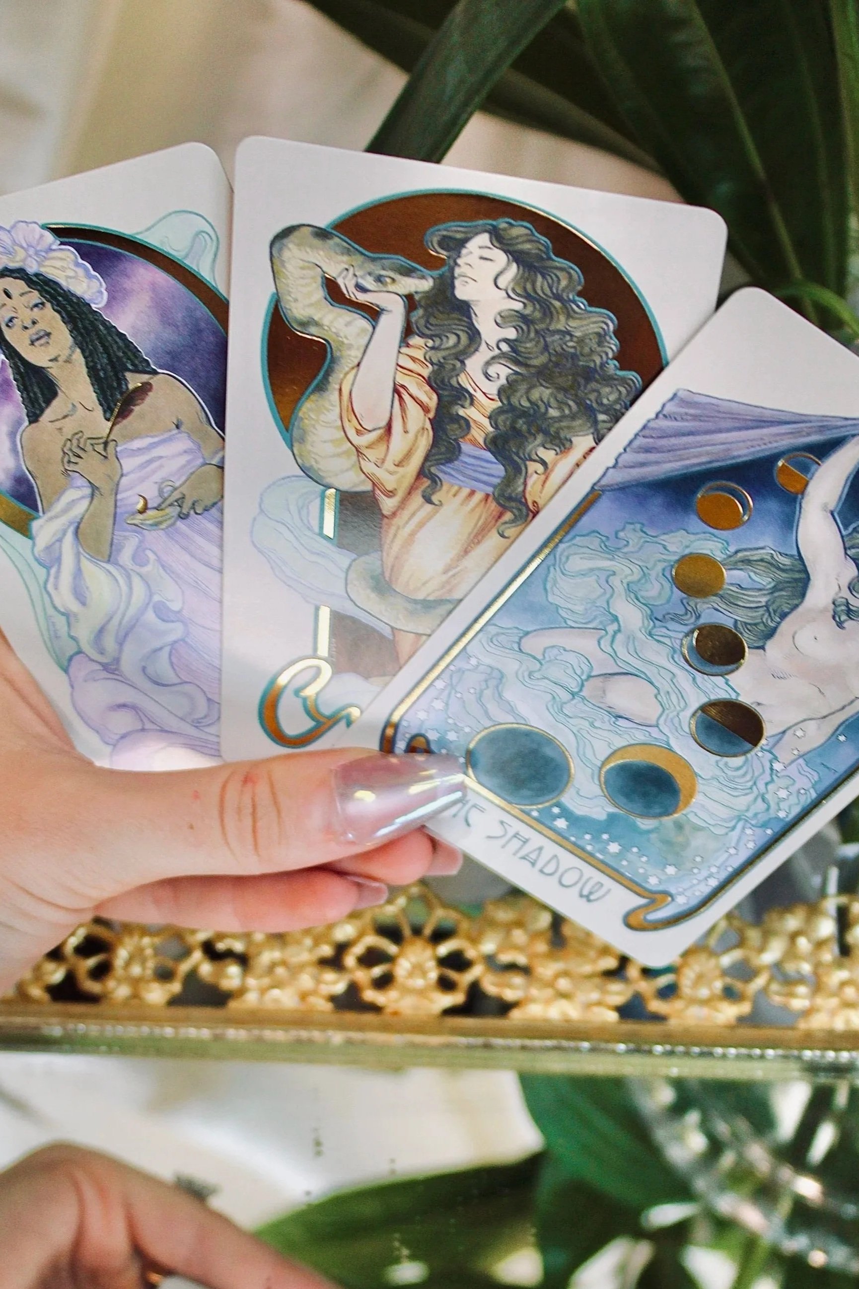 Close-up of tarot cards featuring artistic illustrations of women with long curly hair, one with a snake and the other with water and clouds, held in a person's hand. Background includes green leaves and a decorative box.