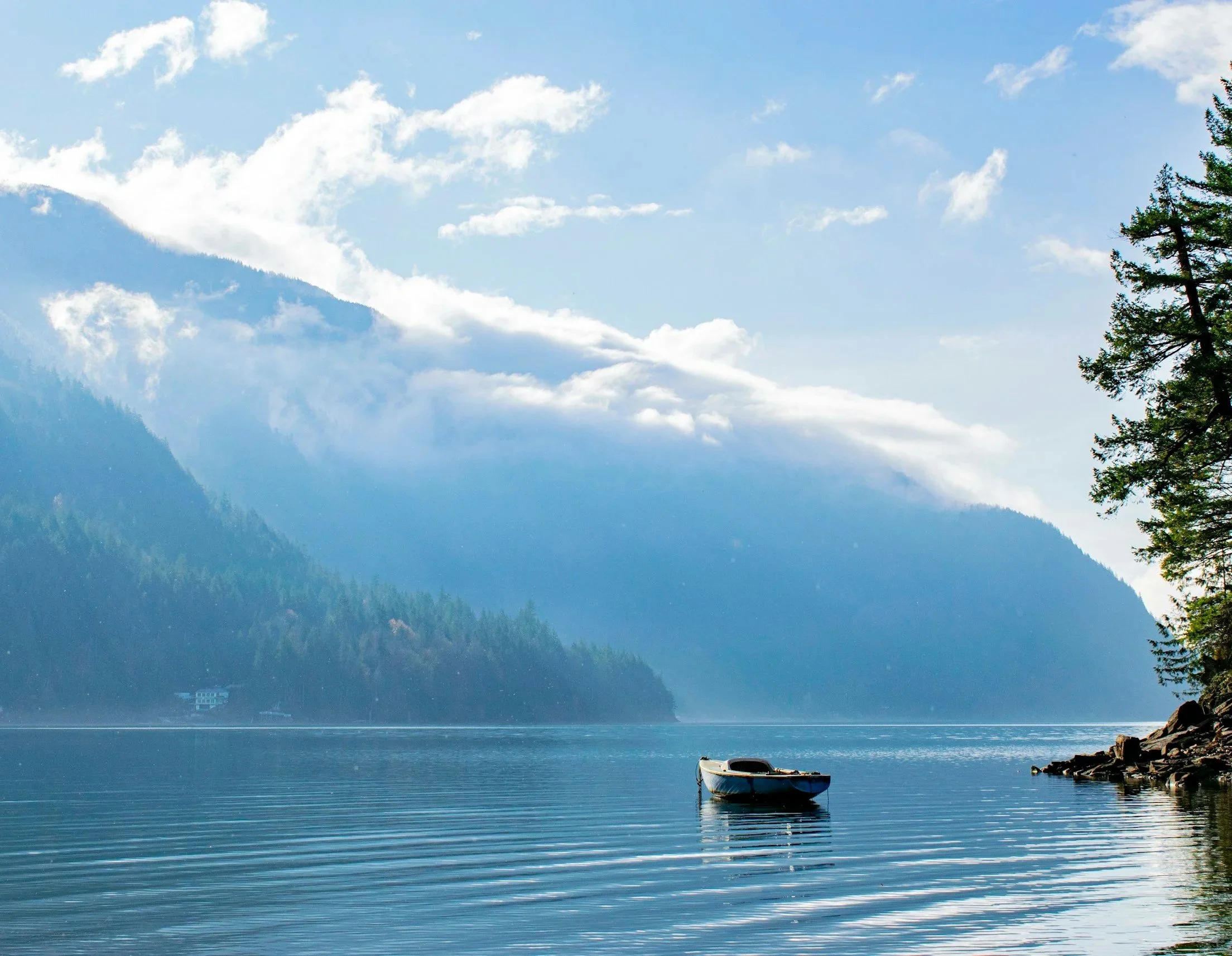 A calm lake with a small boat floating near the rocky shoreline and lush green trees on the right. In the background, mountain hills are partially covered by clouds under a partly cloudy blue sky.