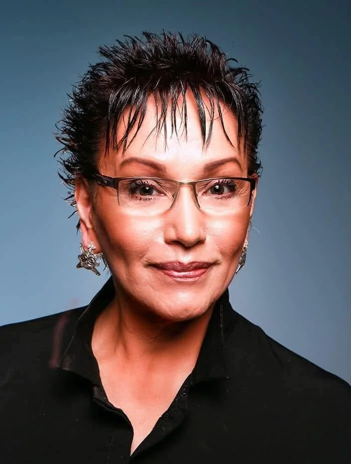 A close-up portrait of a middle-aged woman with short, spiky dark hair, wearing glasses, earrings, and a black top, smiling against a blue background.