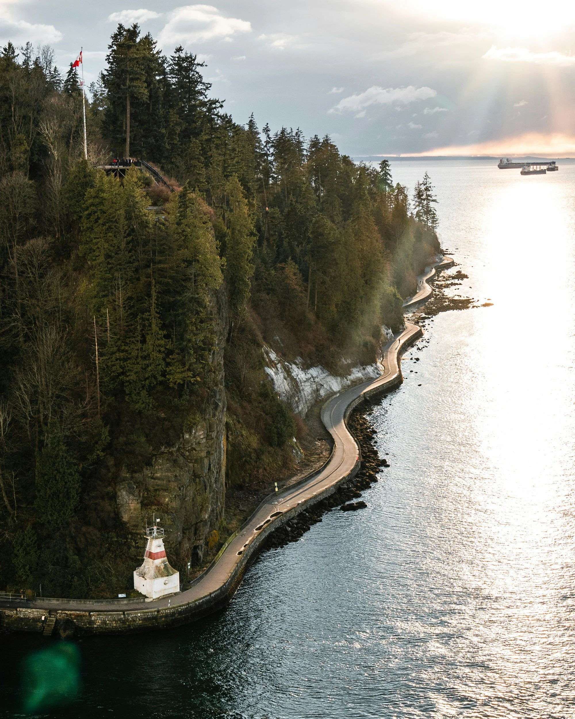 A winding Stanley Park road runs alongside the water, with a lighthouse at the bottom of a forested cliff on the left and cargo ships in the distance on the right. The scene is during a cloudy sunset.