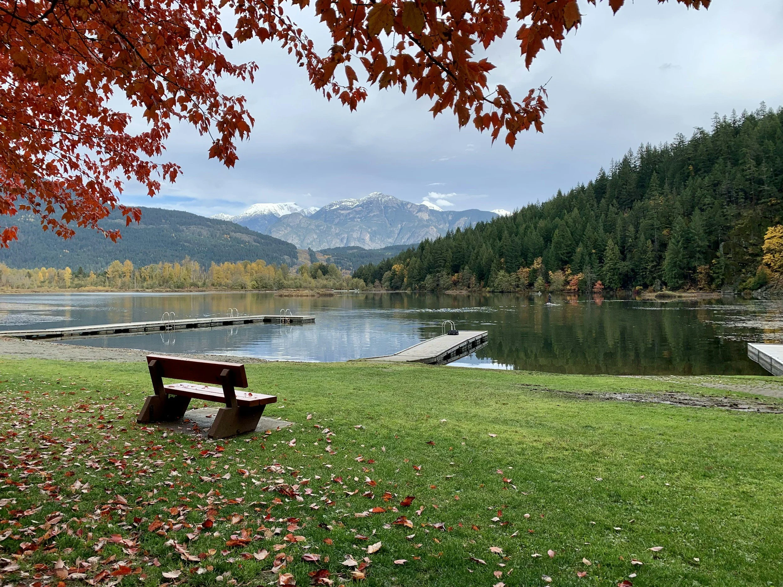A lakeside park scene with a moss-covered bench in the foreground, fallen autumn leaves scattered on green grass, calm lake waters, lush green and yellow trees, mountains with snow caps in the distance, and a partly cloudy sky.