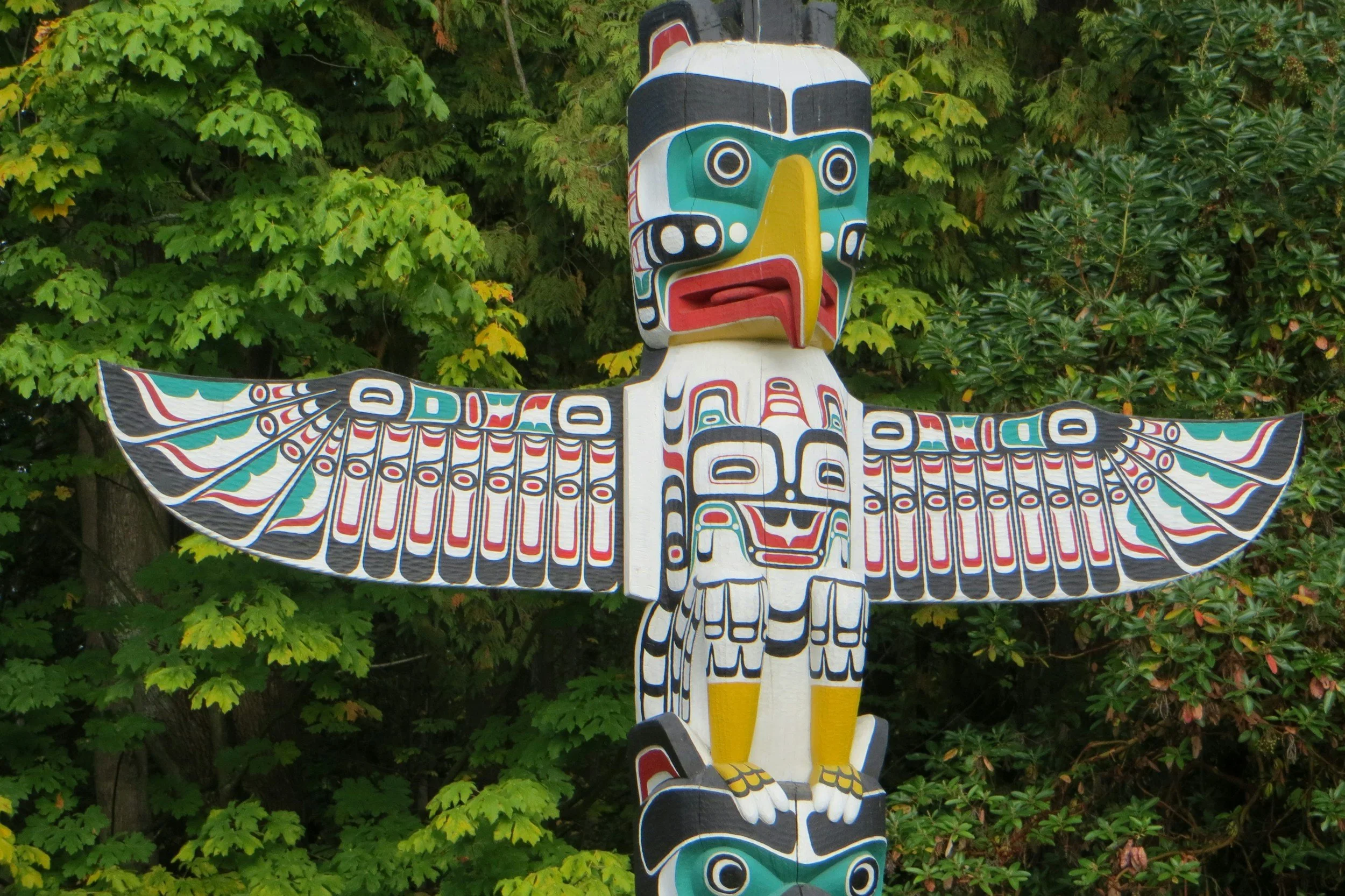 Colorful carved totem pole featuring an eagle and other figures, set against green leafy background.