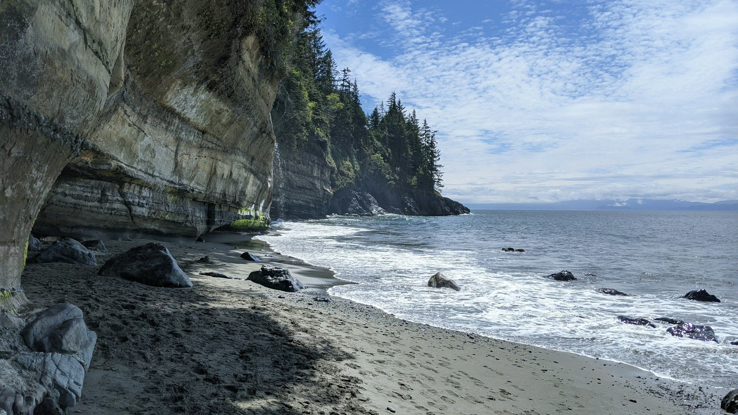 A scenic coastline with large cliffs covered in trees on the left, a sandy beach with rocks at the shoreline, and calm ocean waters extending into the horizon under a partly cloudy sky.