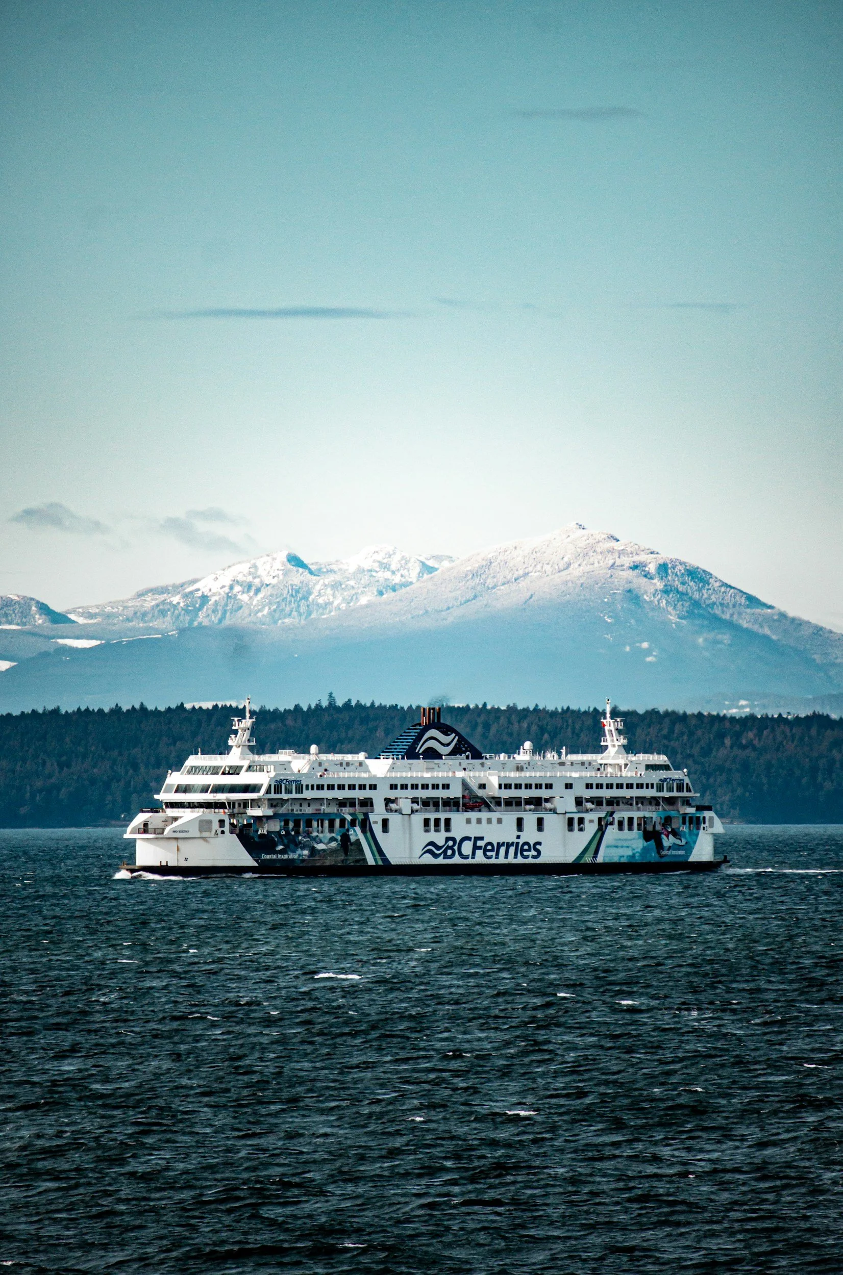 A large white BC Ferries ship sailing in dark blue waters with snow-capped mountains in the background and a clear sky overhead.