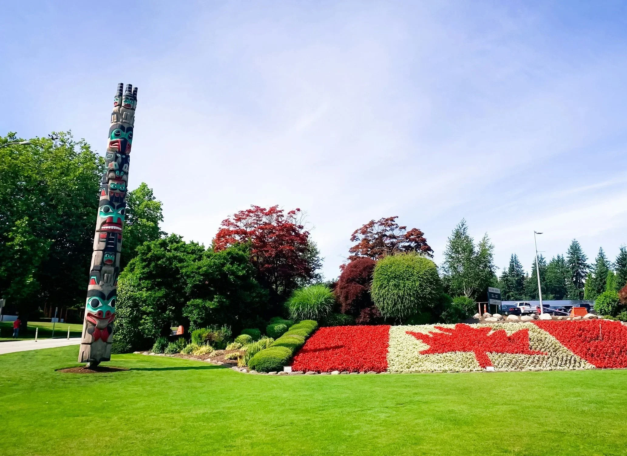 A decorated totem pole surrounded by green trees, flowering bushes, and a well-maintained lawn with a red and white floral display, under a clear blue sky.