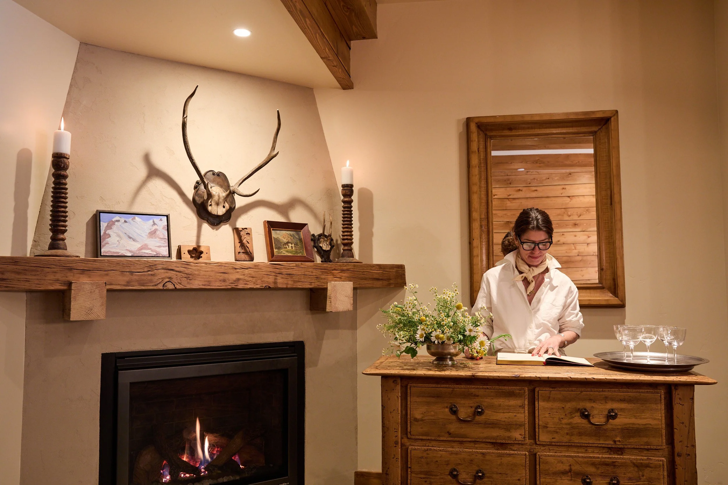 Interior of a cozy room with a fireplace, decorating items including a deer antler mount, a painting, and candles. A woman with glasses is arranging flowers on a wooden table.
