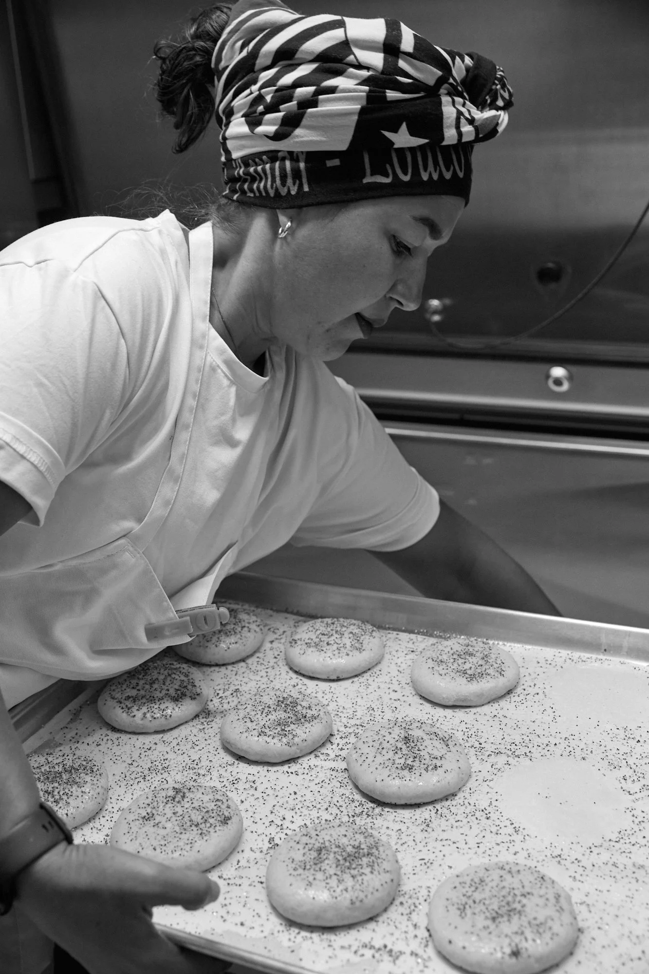 A female cook in a white shirt is placing round cookies or bread rolls on a baking tray in a kitchen. The background is a kitchen environment.
