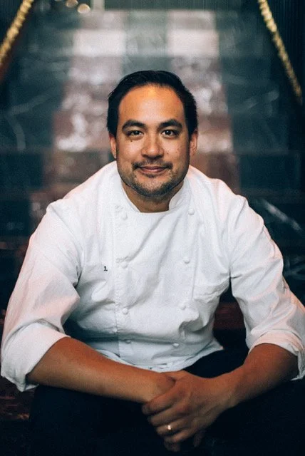 A male chef in a white chef coat sitting on stairs with a dark background, smiling at the camera.