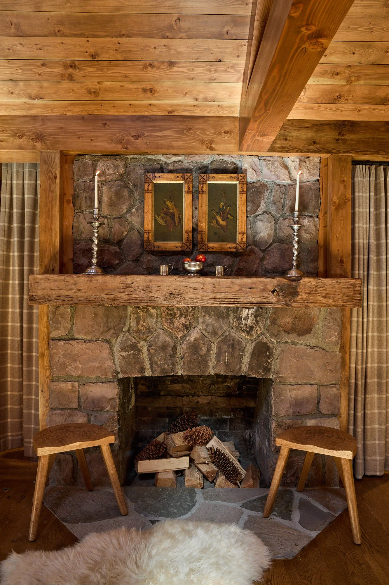 A rustic living room featuring a stone fireplace with wooden beams and two chairs on either side. The fireplace has a wooden mantle with two silver candlesticks holding lit candles, a bowl of apples, and two framed bird illustrations above. Firewood 
