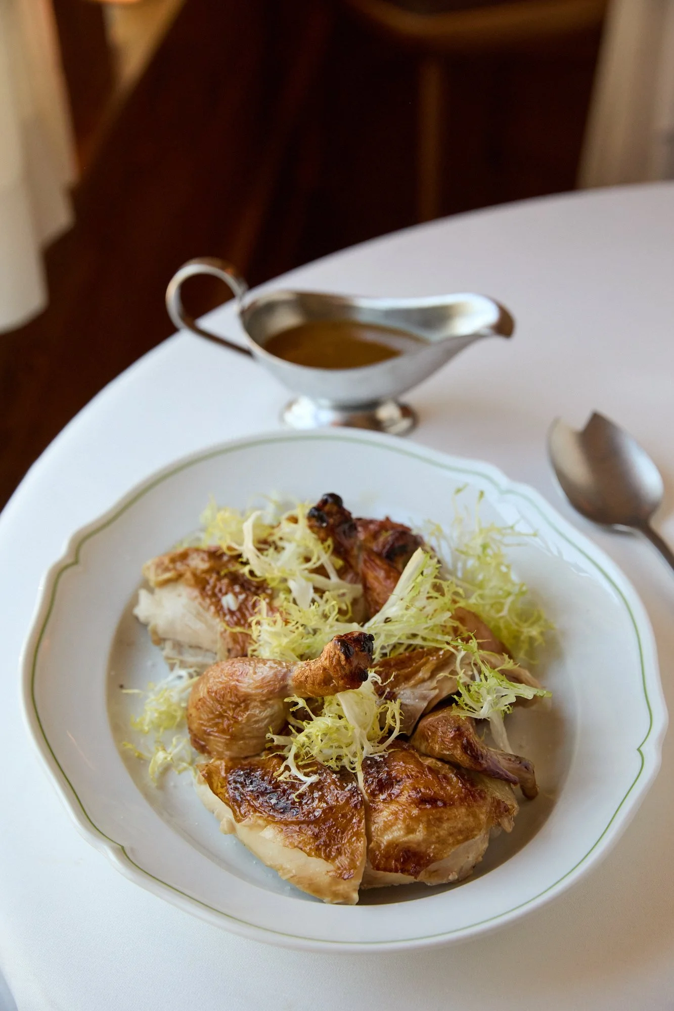 A white plate with roasted chicken pieces topped with shredded lettuce, on a white tablecloth. In the background, there is a silver gravy boat on the table, and a spoon nearby.
