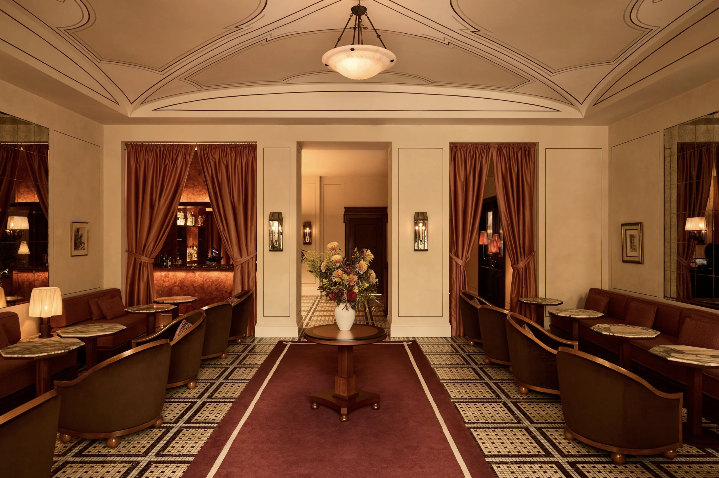 Elegant lounge area with a central table holding a large floral arrangement, surrounded by brown chairs, with a separate seating area behind loaded with warm lighting, curtains, and vintage-style decor.