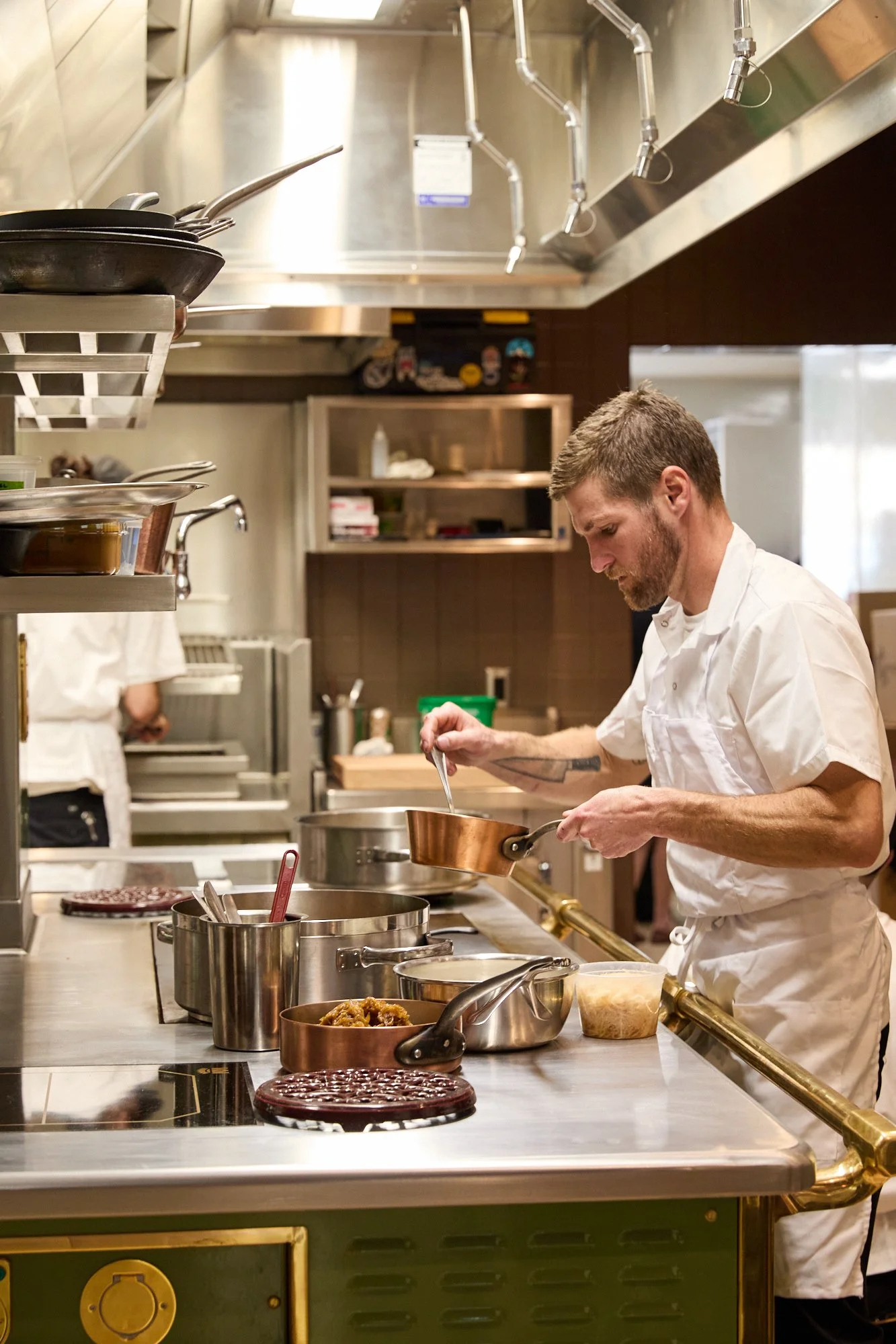 A male chef in a white uniform preparing food in a professional kitchen, surrounded by pots, pans, and cooking utensils.