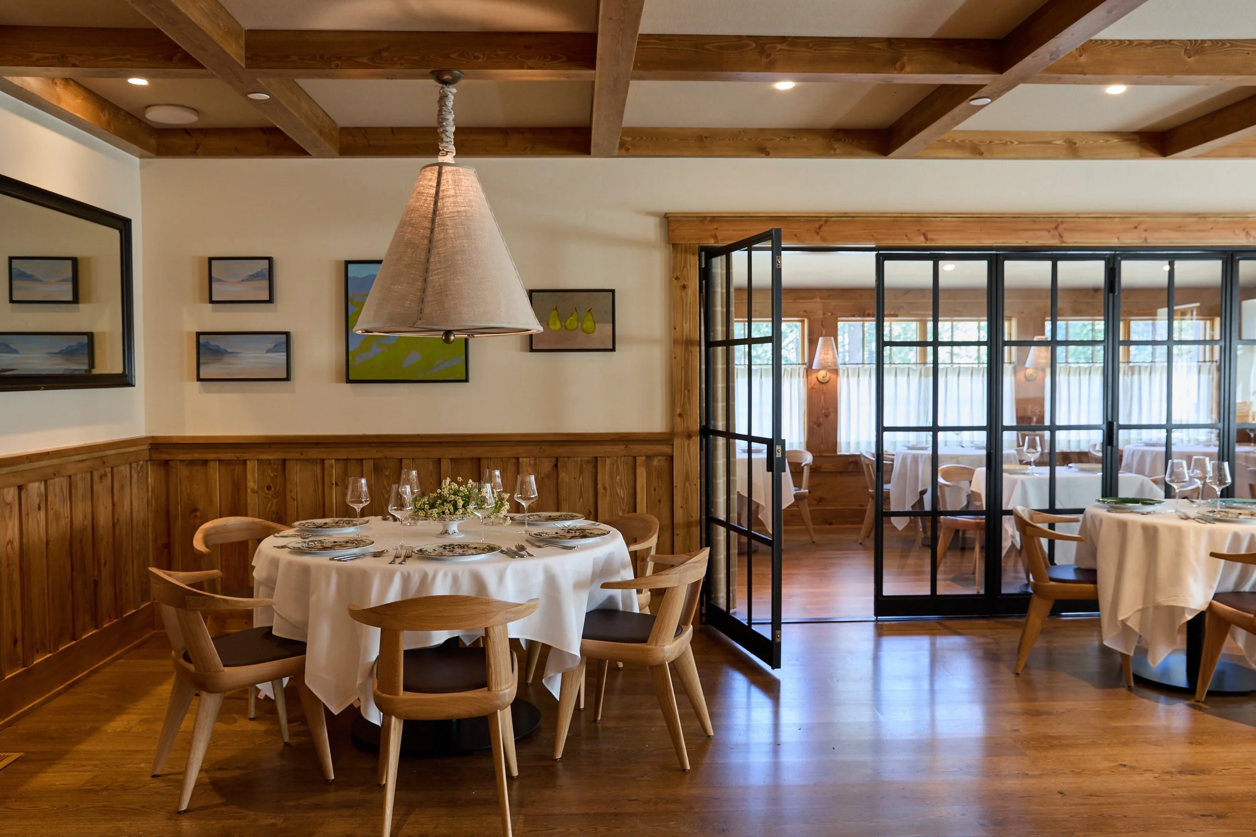 A restaurant dining room with round tables covered with white tablecloths, wooden chairs, and elegant place settings with wine glasses. The room features wooden beams on the ceiling, framed artwork on the walls, and large glass doors with curtains leading to another dining area with more tables and chairs.
