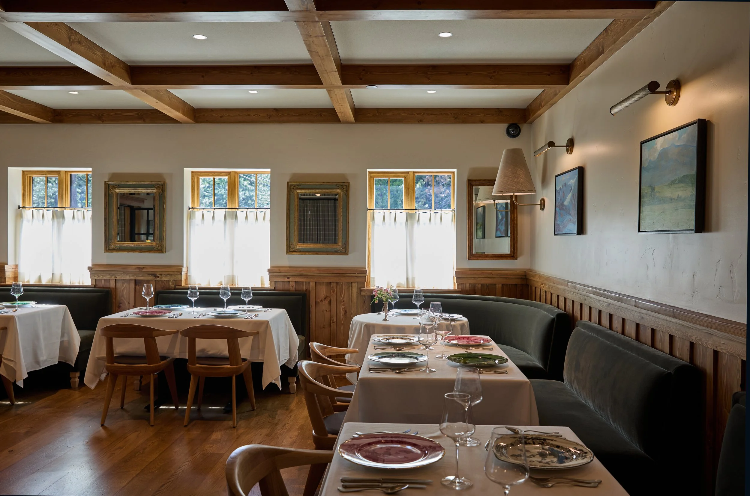 Interior of a restaurant with white tablecloths, wine glasses, and plates on the tables, wooden chairs, and decorated walls with framed artwork and a mirror, with windows allowing natural light in.