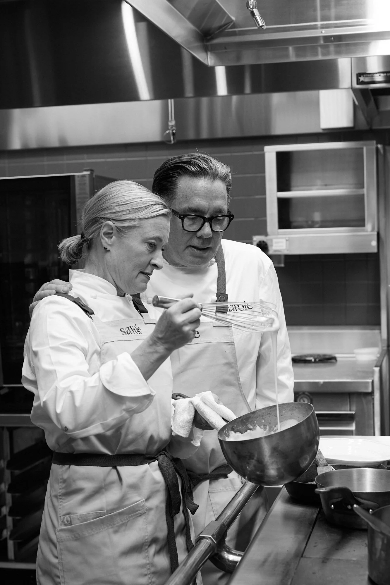 Two chefs in a commercial kitchen preparing food. The woman is holding a whisk and looking into a bowl, while the man observes. Both are dressed in chef uniforms.