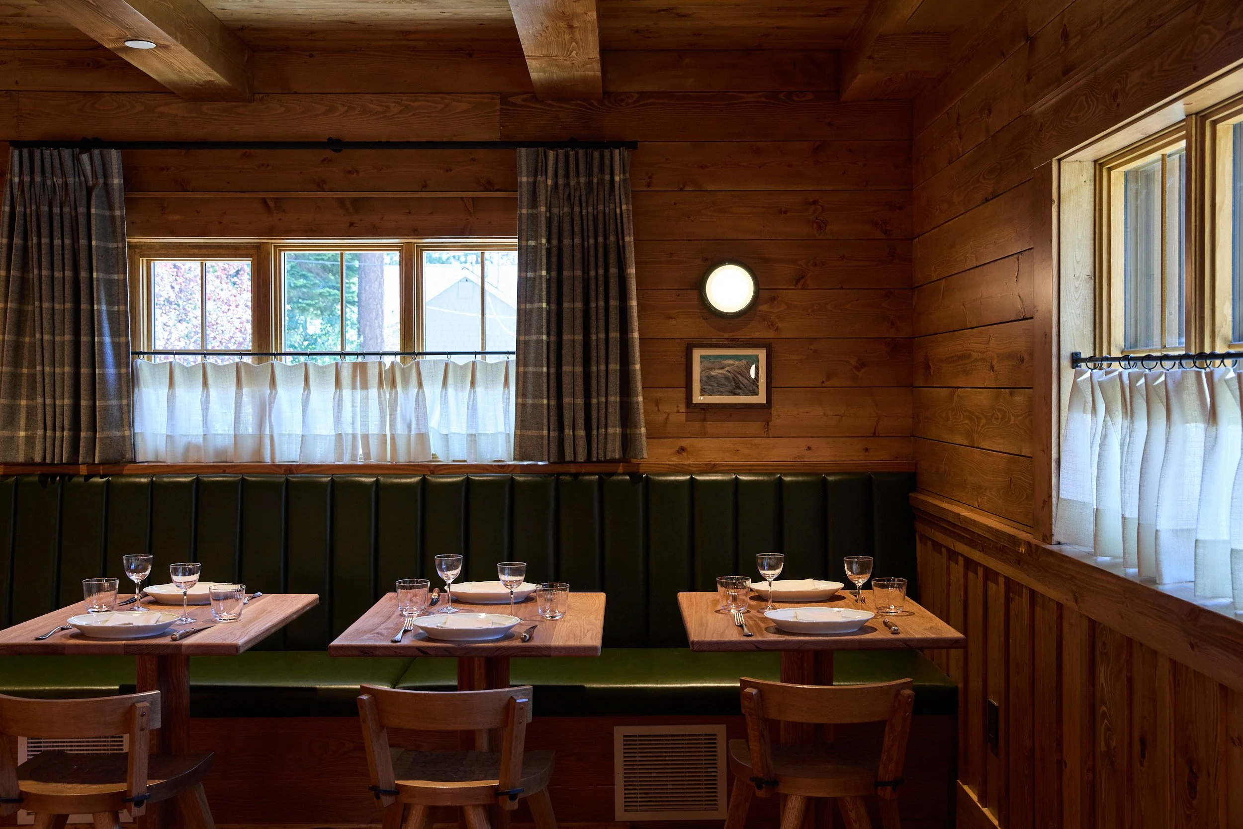 A cozy dining area with wooden walls and windows, set with plates, glasses, and silverware on wooden tables, featuring green booth seating and chairs.