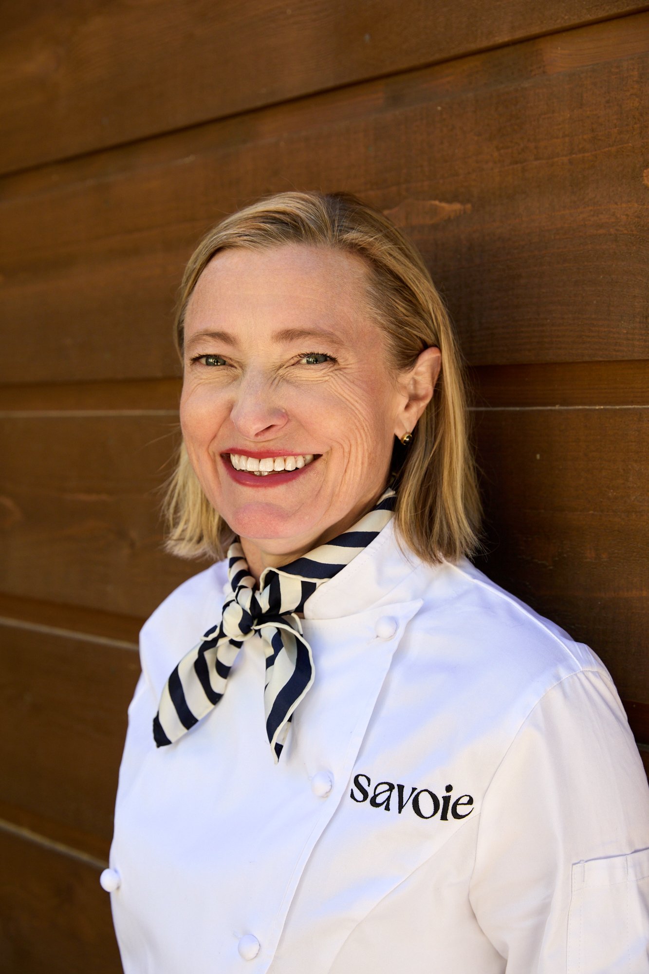 Close-up of a smiling woman in a white chef's coat with 'savoie' embroidered, wearing a black and white striped neckerchief, standing against a wooden background.