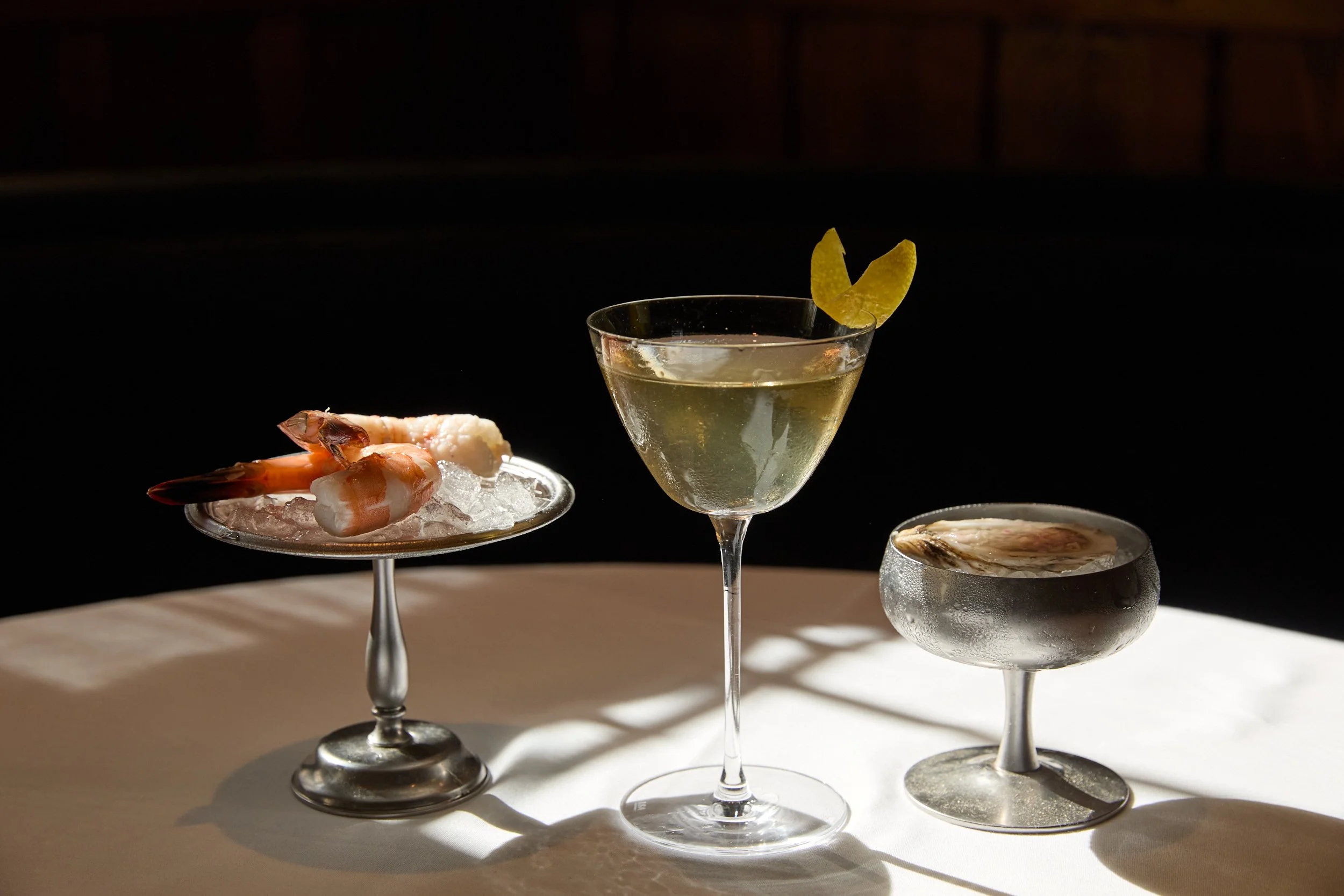 A glass of cocktail with a lemon twist garnish, a plate of shrimp on ice, and a bowl of oysters on ice, all arranged on a white surface against a dark background.