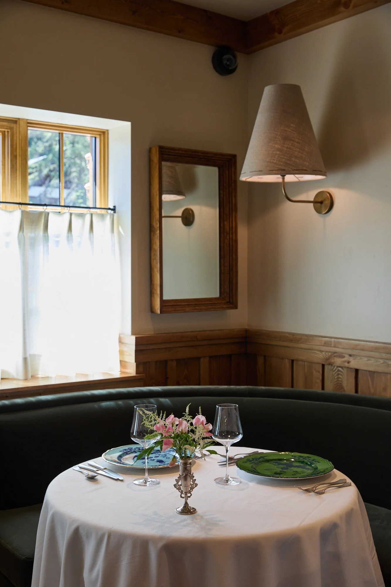 A round dining table set with a white tablecloth, a floral centerpiece, two wine glasses, and plates, in a cozy room with a window, a mirror, and a wall-mounted lamp.
