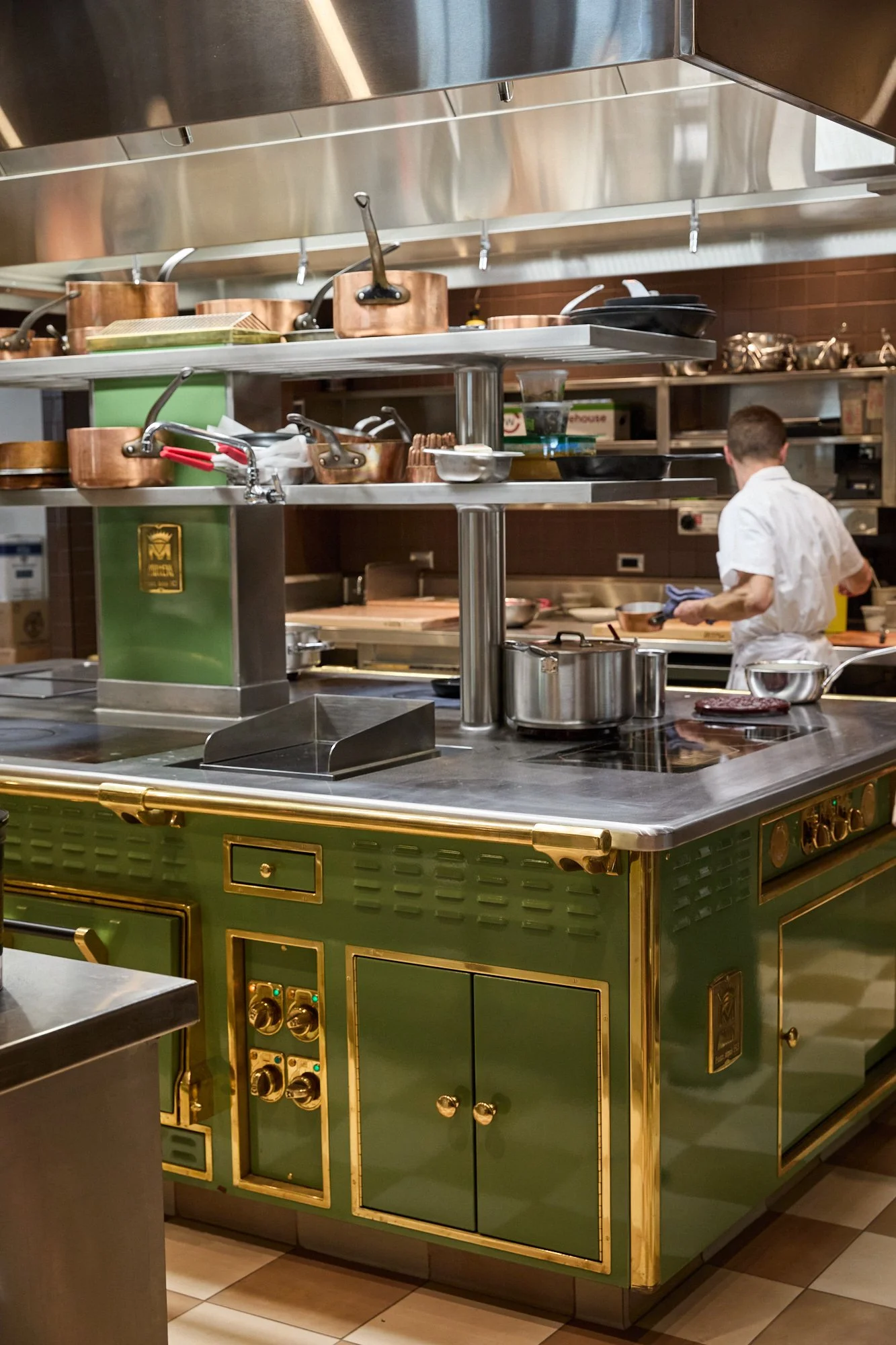 A professional kitchen with a chef working behind a green and gold cooking station, surrounded by various pots and utensils.