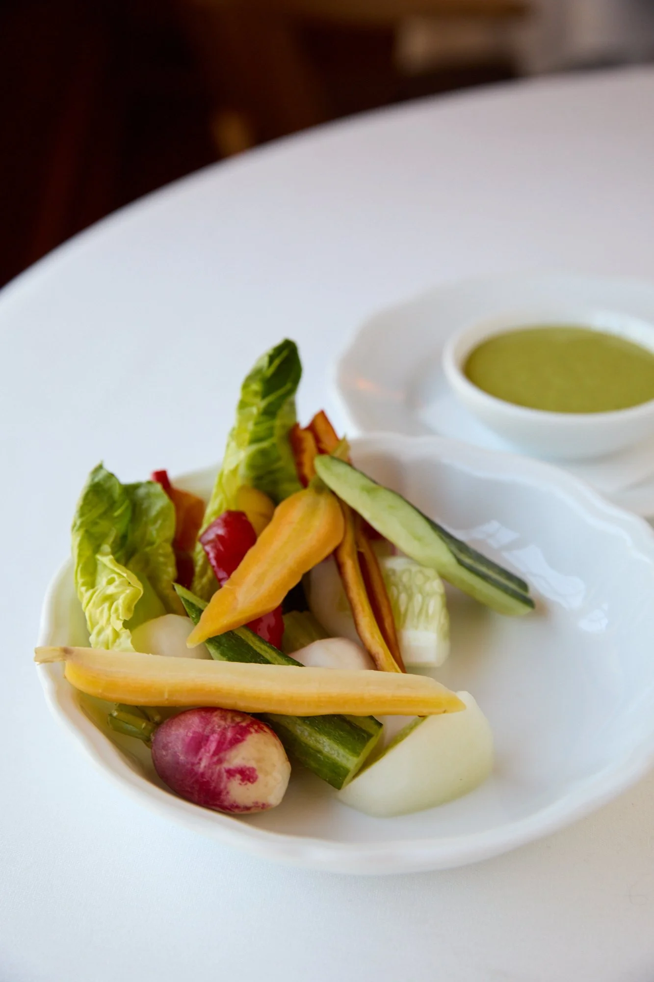 A white bowl filled with colorful mixed vegetables including radishes, carrots, celery, and lettuce, served alongside a small dish of green sauce or dressing on a white plate.