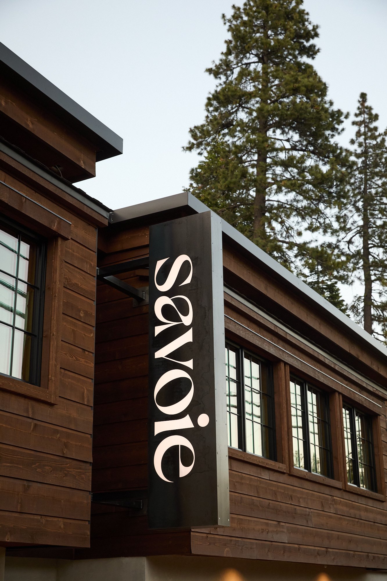 Close-up of a black sign with white text attached to a wooden building, with the word 'Vogue' visible, and tall trees in the background.