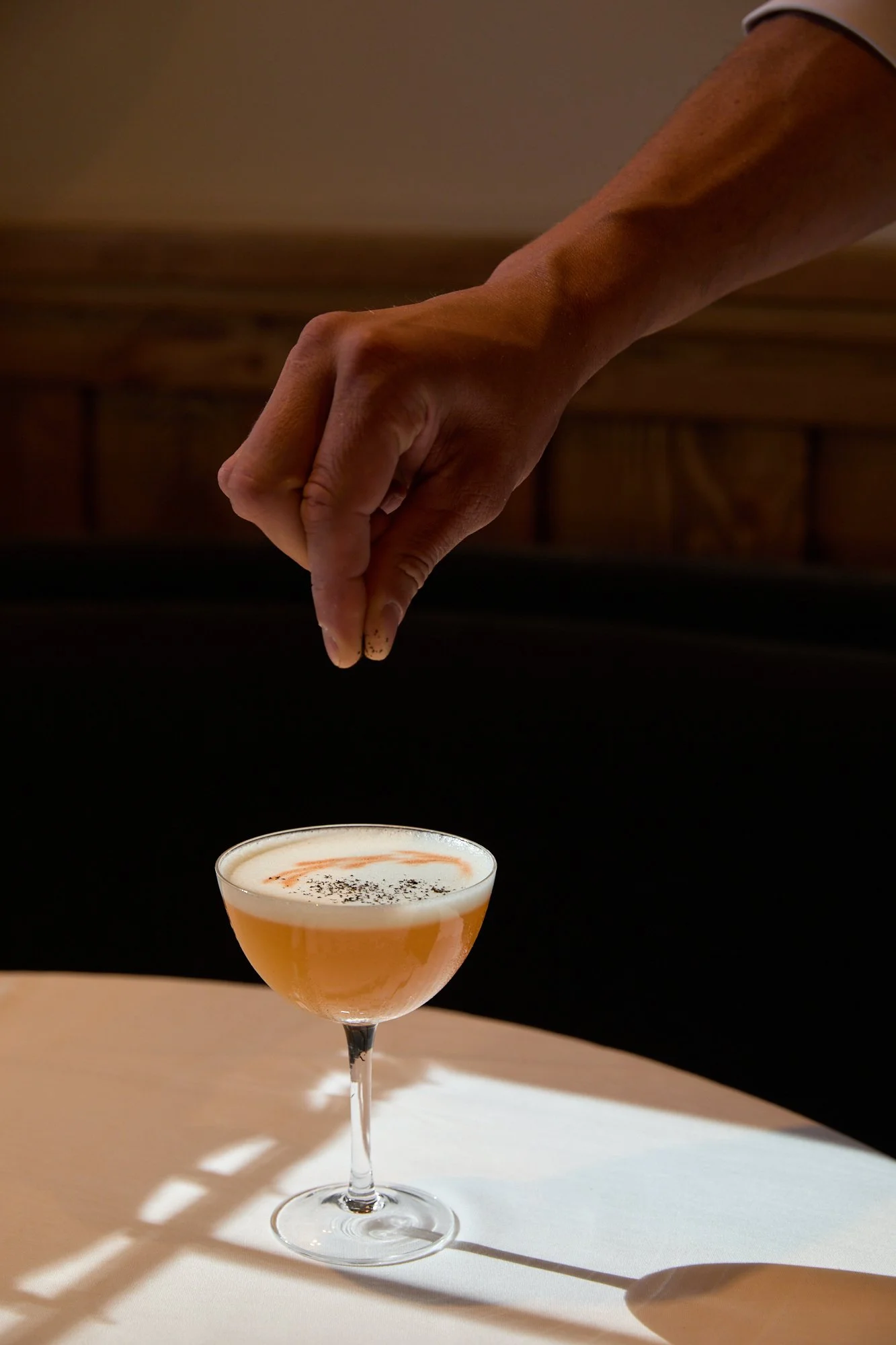 A person is sprinkling black pepper onto a peach-colored cocktail in a coupe glass on a white surface, with shadows cast on the table.