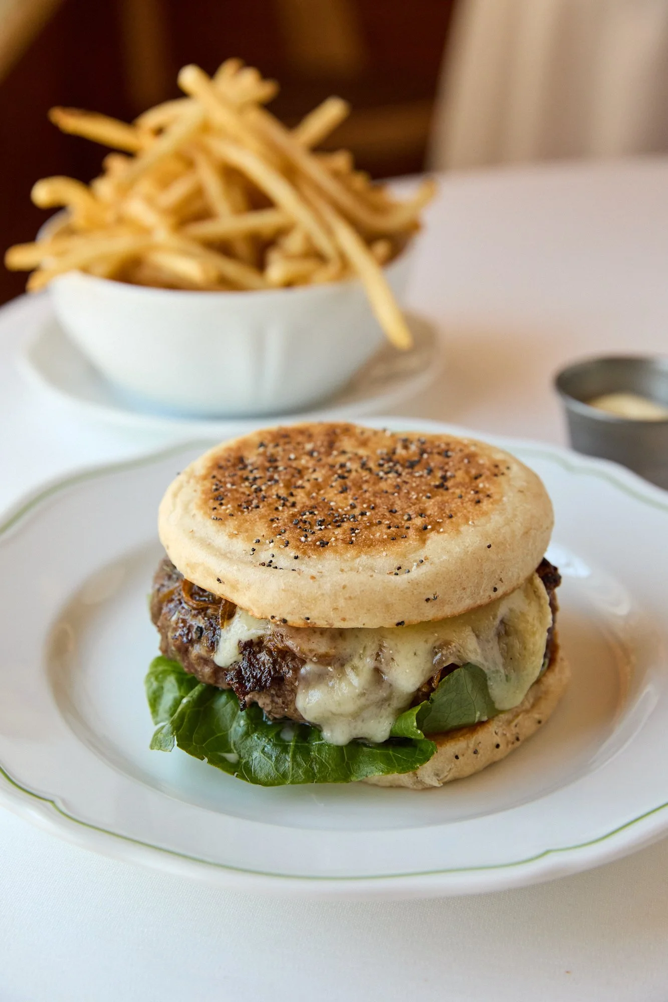 A cheeseburger with lettuce, melted cheese, and a toasted bun on a white plate, with a side of French fries in a white bowl and a ramekin of aioli in the background.