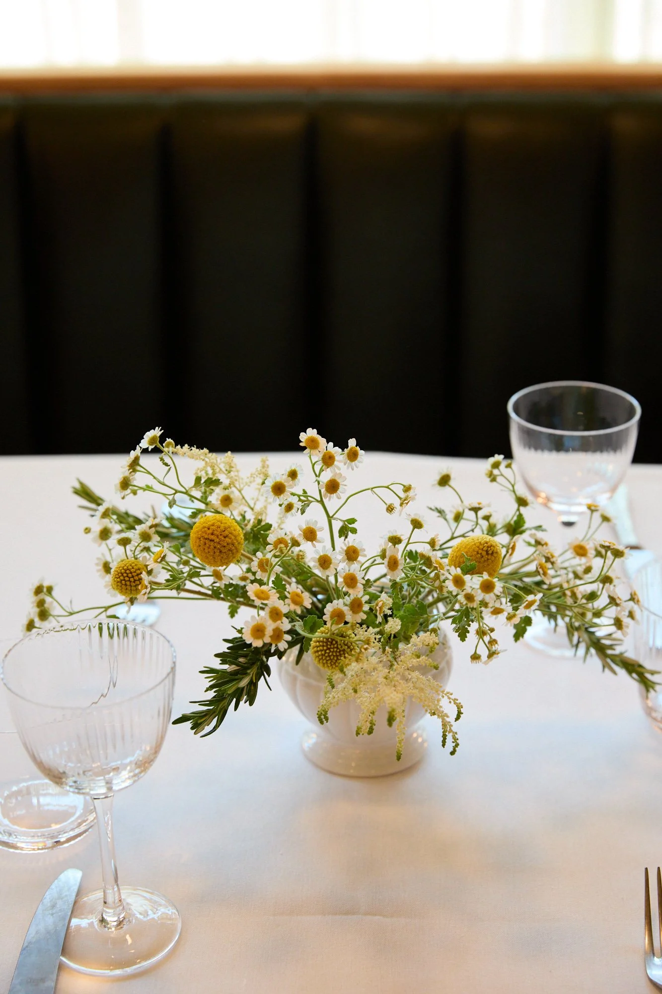 A table setup with a floral centerpiece including white and yellow flowers in a white vase, empty crystal glasses, a knife, and a fork on a white tablecloth, with a black padded headboard or wall in the background.