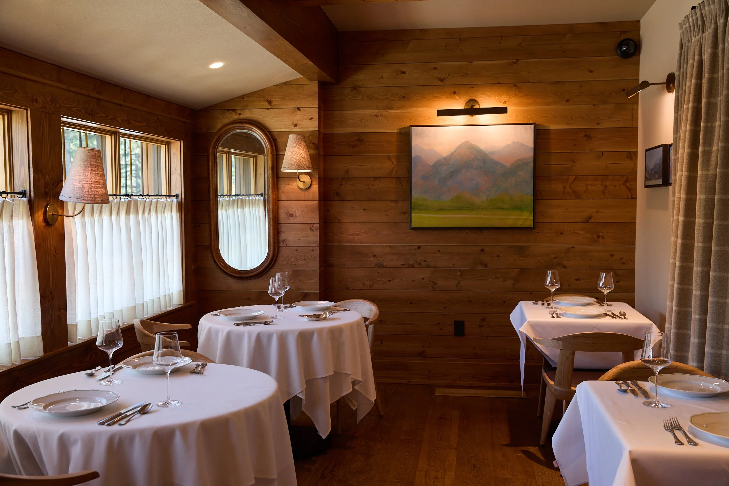 A cozy dining room with wooden walls and large windows with cream curtains. The room has round tables covered with white tablecloths, set with plates, silverware, and wine glasses. There are wall-mounted lamps and a landscape painting on the wall.