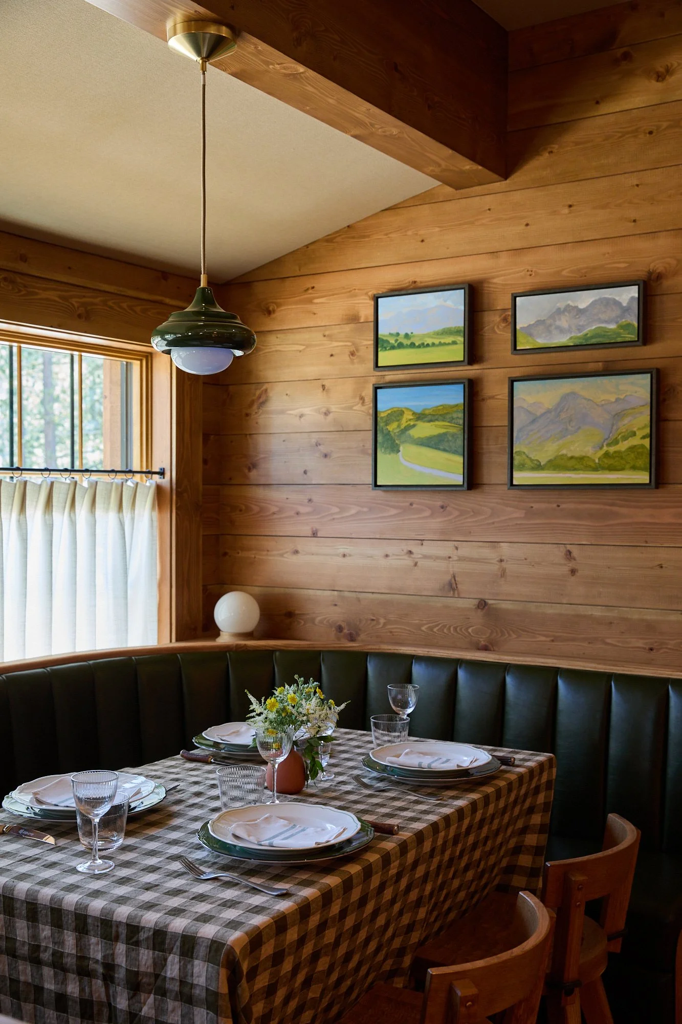 A cozy dining room with wood-paneled walls and a window with curtains. There is a black leather booth around a checkered tablecloth with place settings, glasses, silverware, and a flower vase with yellow flowers. A hanging pendant light is above the 