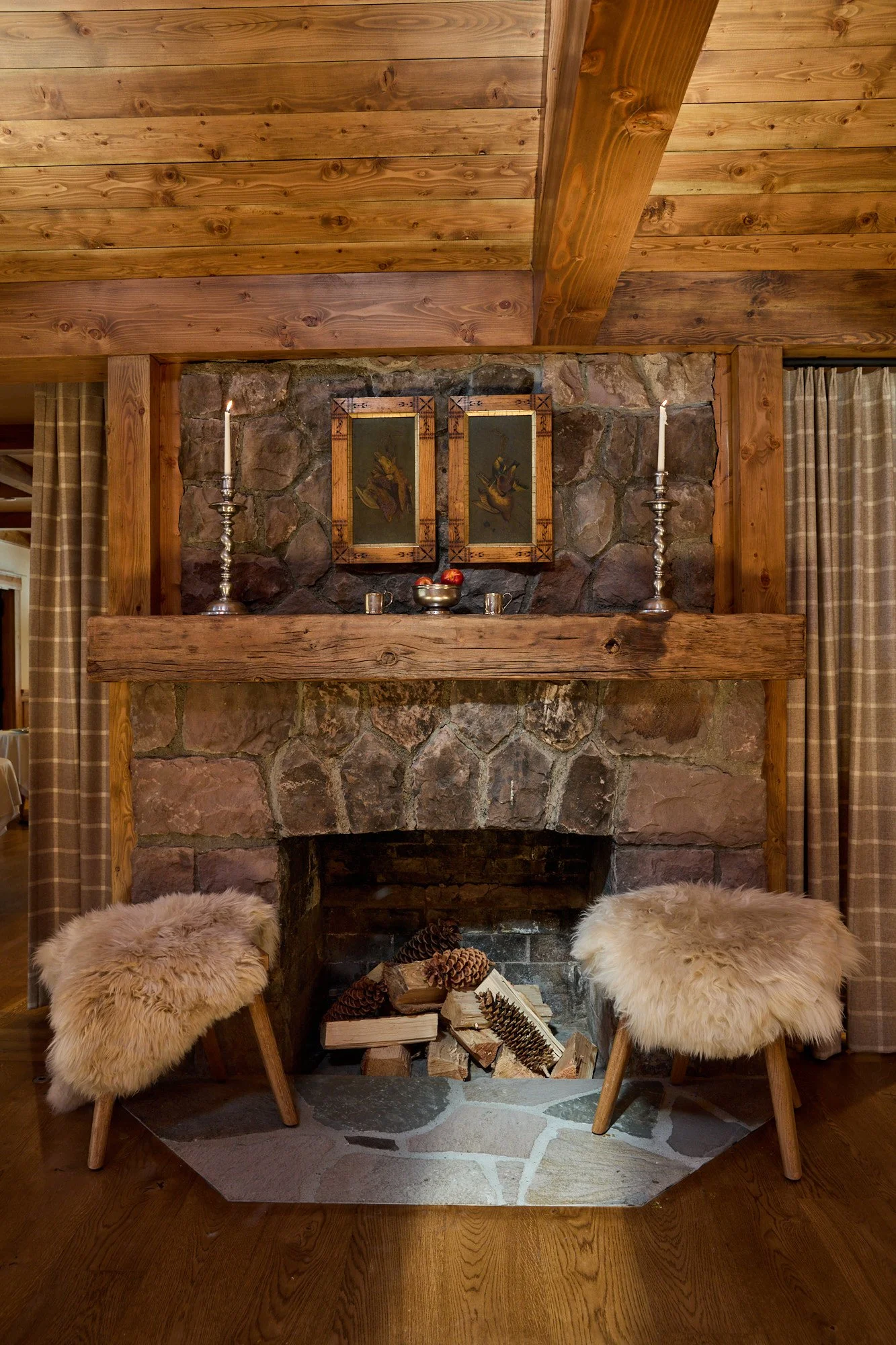 A rustic living room feature with a stone fireplace, two wooden chairs with fluffy sheepskin covers, a wooden mantle with candles and framed artwork, and a pile of firewood inside the fireplace. The ceiling and surrounding walls are made of wood, and curtains are visible on either side.