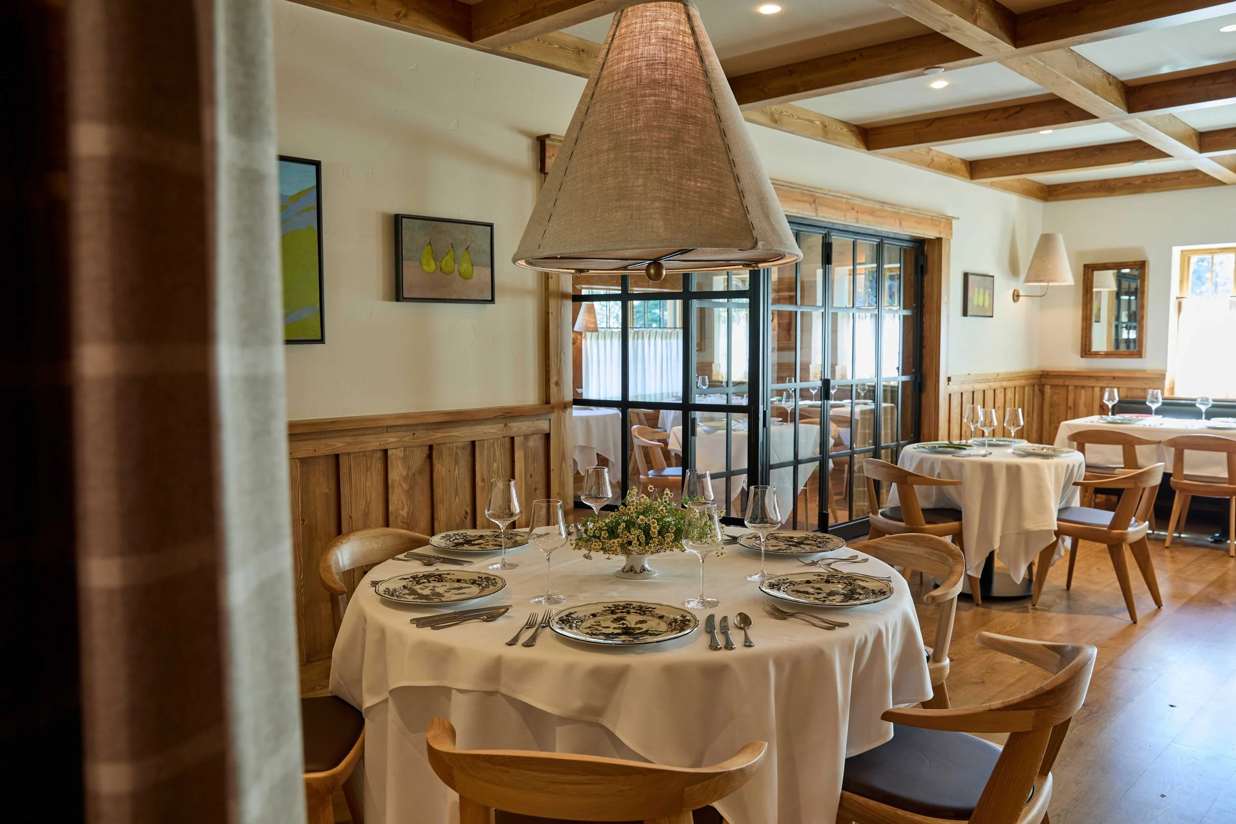 Interior of a restaurant with a round table set for dining, featuring a white tablecloth, floral dinner plates, wine glasses, and silverware. The room has wooden beam ceilings, framed artwork on the walls, and a large window letting in natural light. There are additional tables with similar settings and wooden chairs around the room.