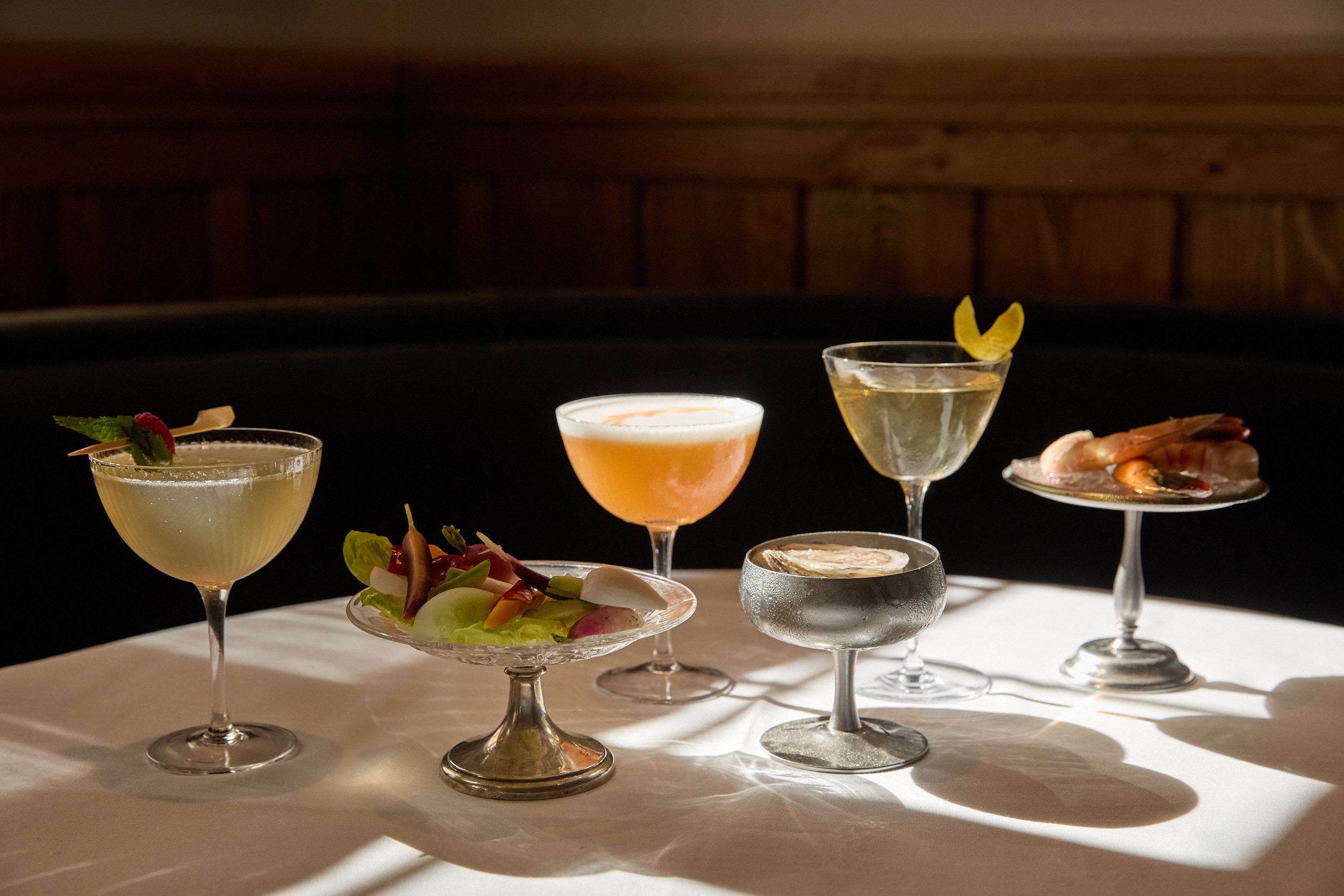 Assorted cocktails with a shrimp and oyster appetizer on a table with a dark wood background.