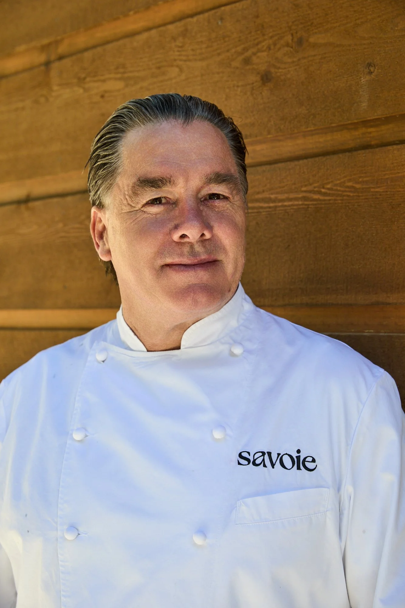 A man in a white chef's coat standing against a wooden background.