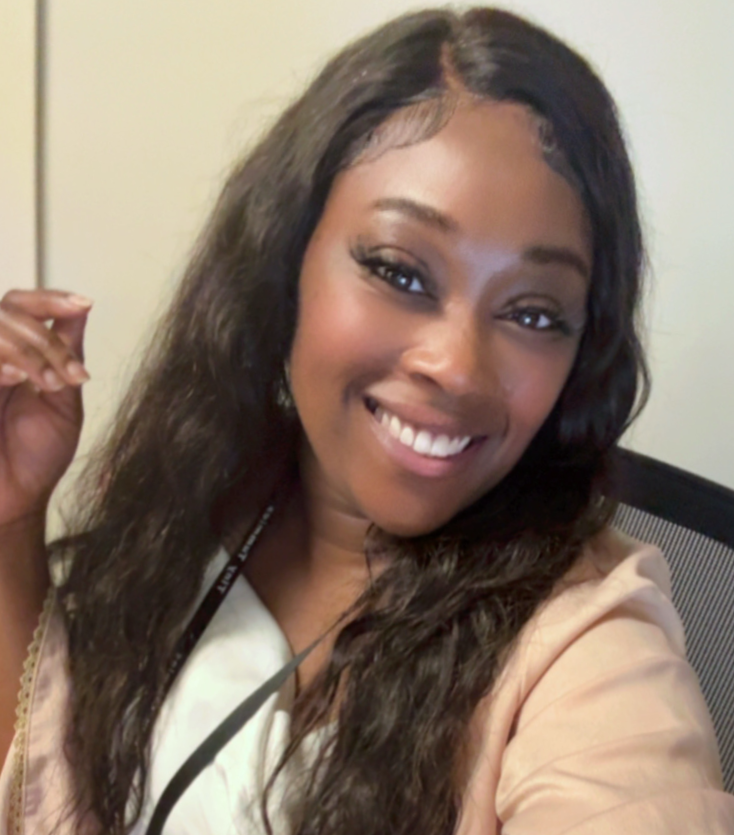 A woman smiling with long wavy hair, wearing a light-colored blouse, sitting in front of a beige wall.