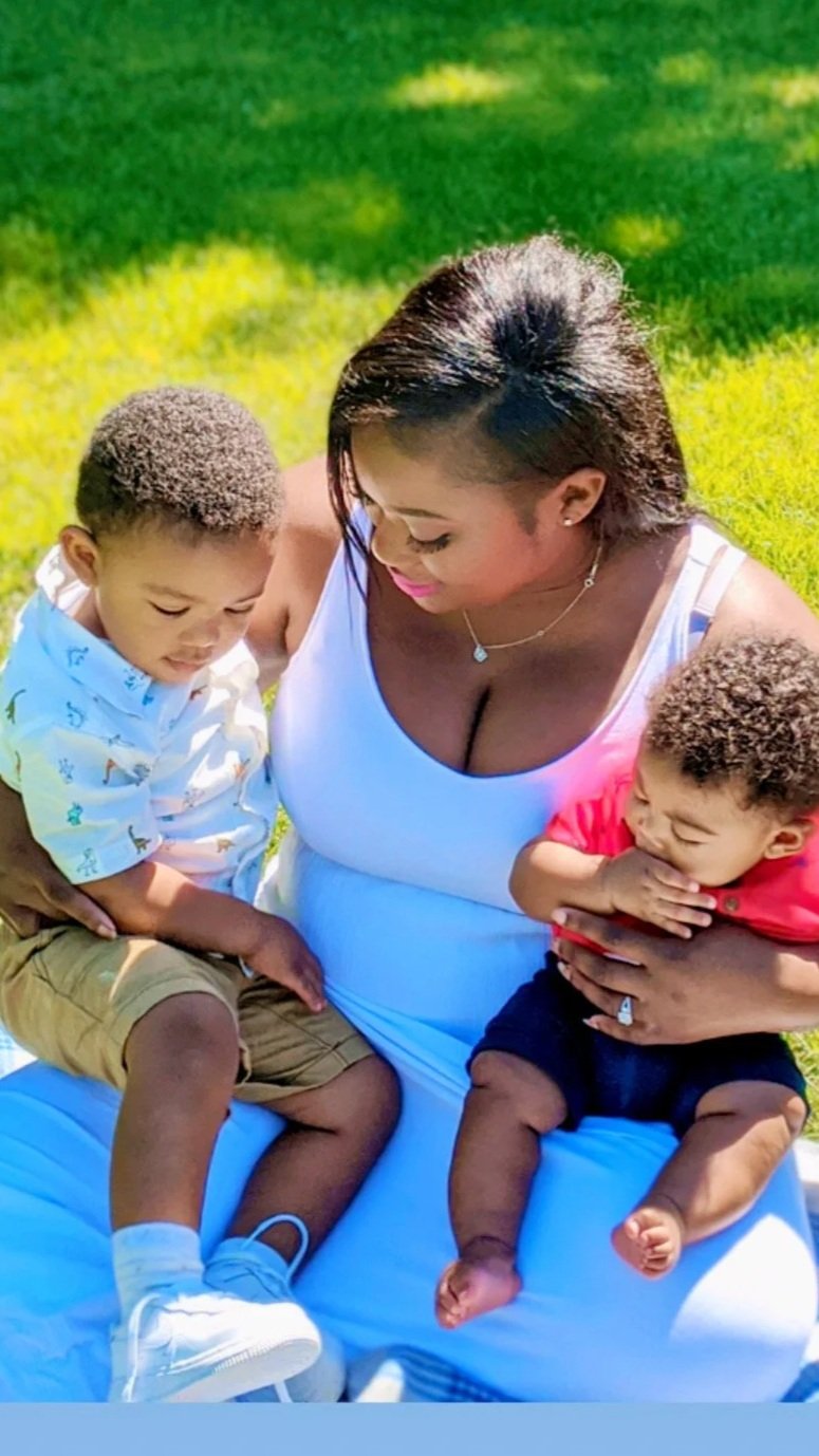 A woman sitting outdoors on grass, holding two young children, one in each arm. She is looking down at the children, who are seated on her lap, against a background of greenery.