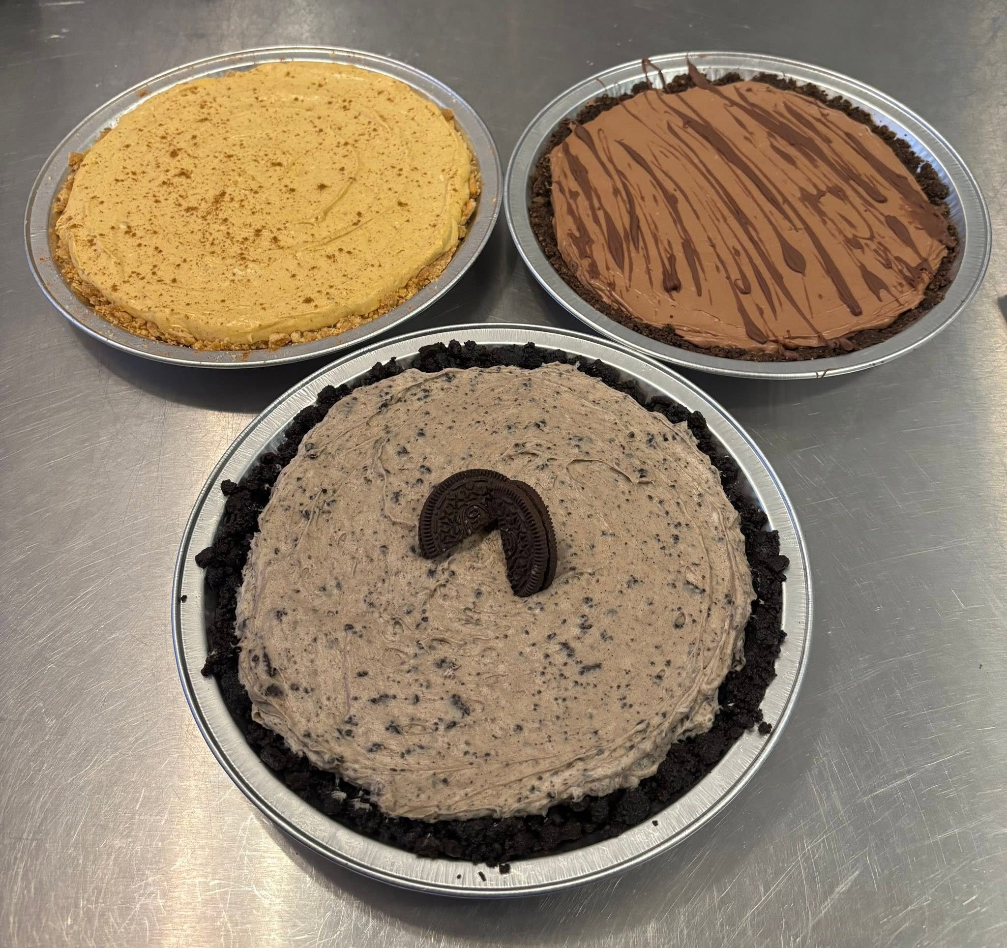 Four different types of slices of pie or cheesecake in aluminum containers, featuring a variety of flavors and toppings, on a metallic surface.