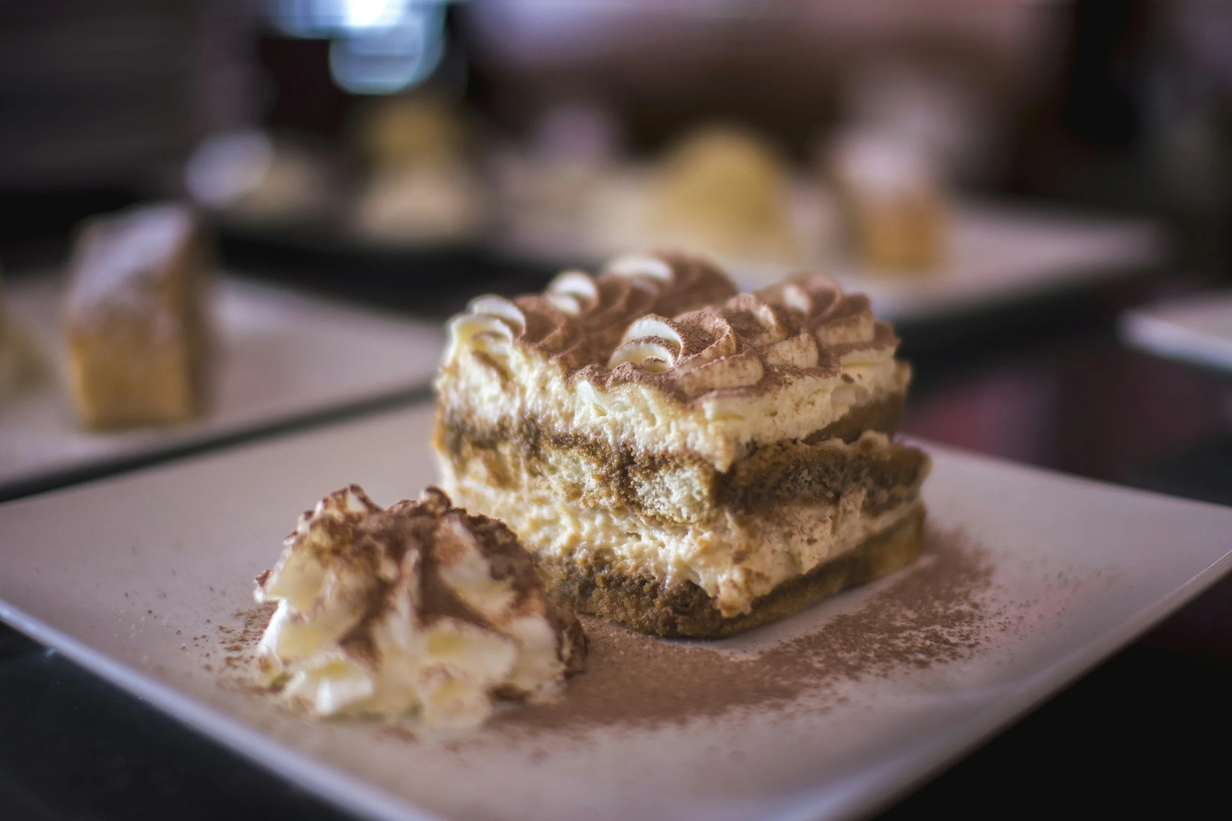 Close-up of a slice of layered cheesecake with chocolate and cream toppings on a white plate, with other desserts blurred in the background.