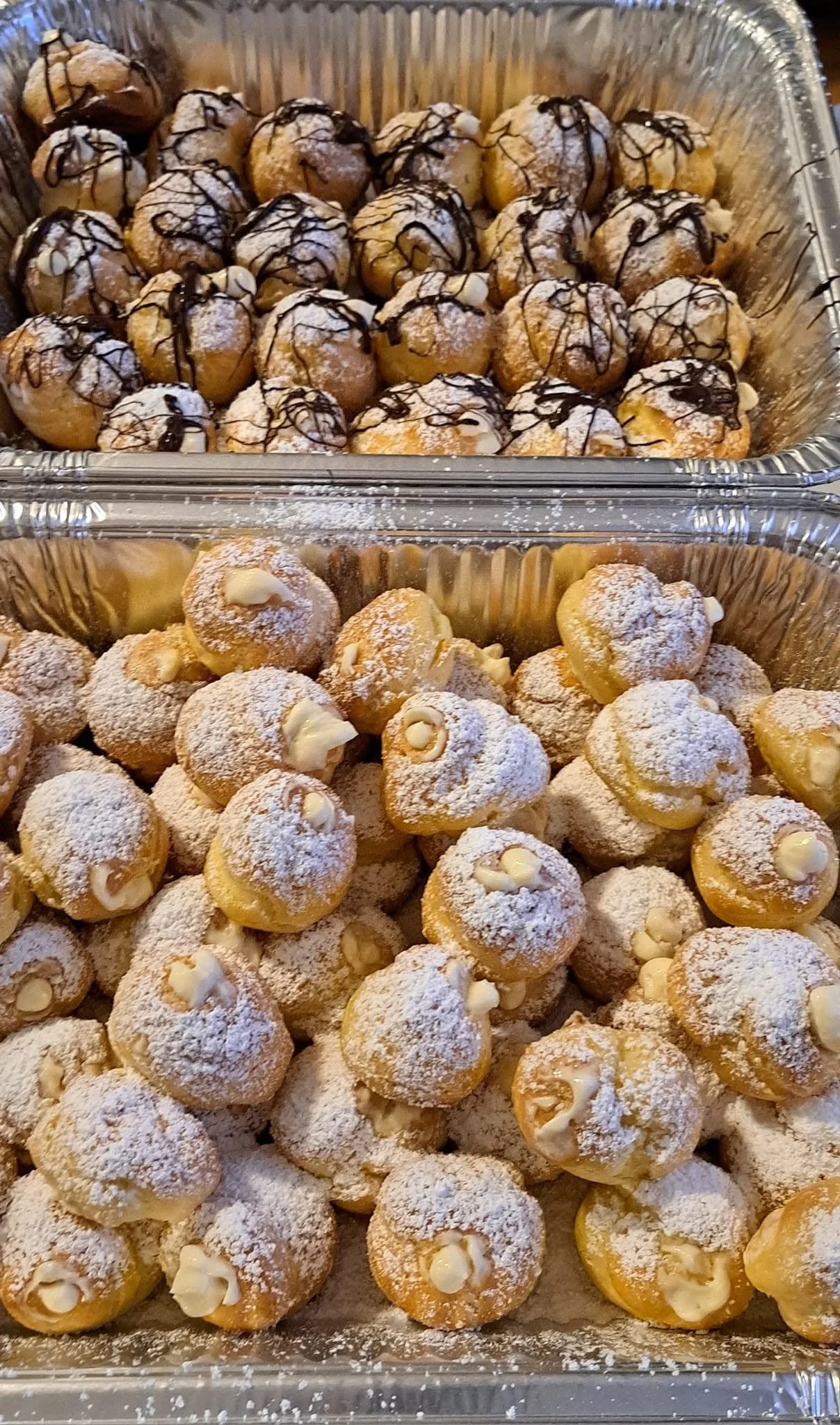 Two aluminum trays filled with assorted cream puffs, dusted with powdered sugar, drizzled with chocolate, and decorated with small chunks of white chocolate.