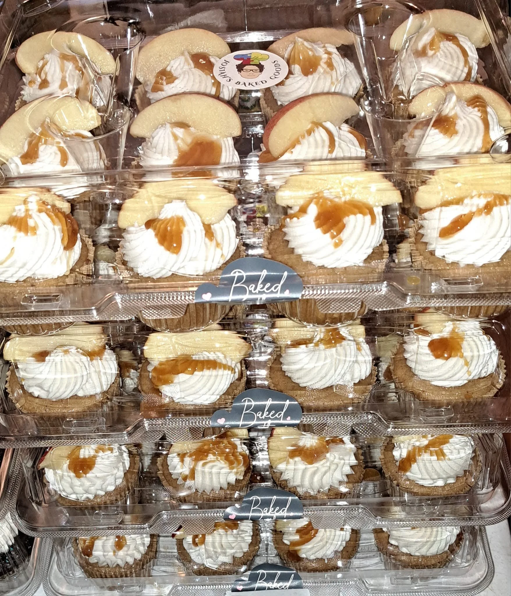 Multiple mini cheesecakes with whipped cream, caramel drizzle, and a slice of apple on top, arranged in a plastic display tray.