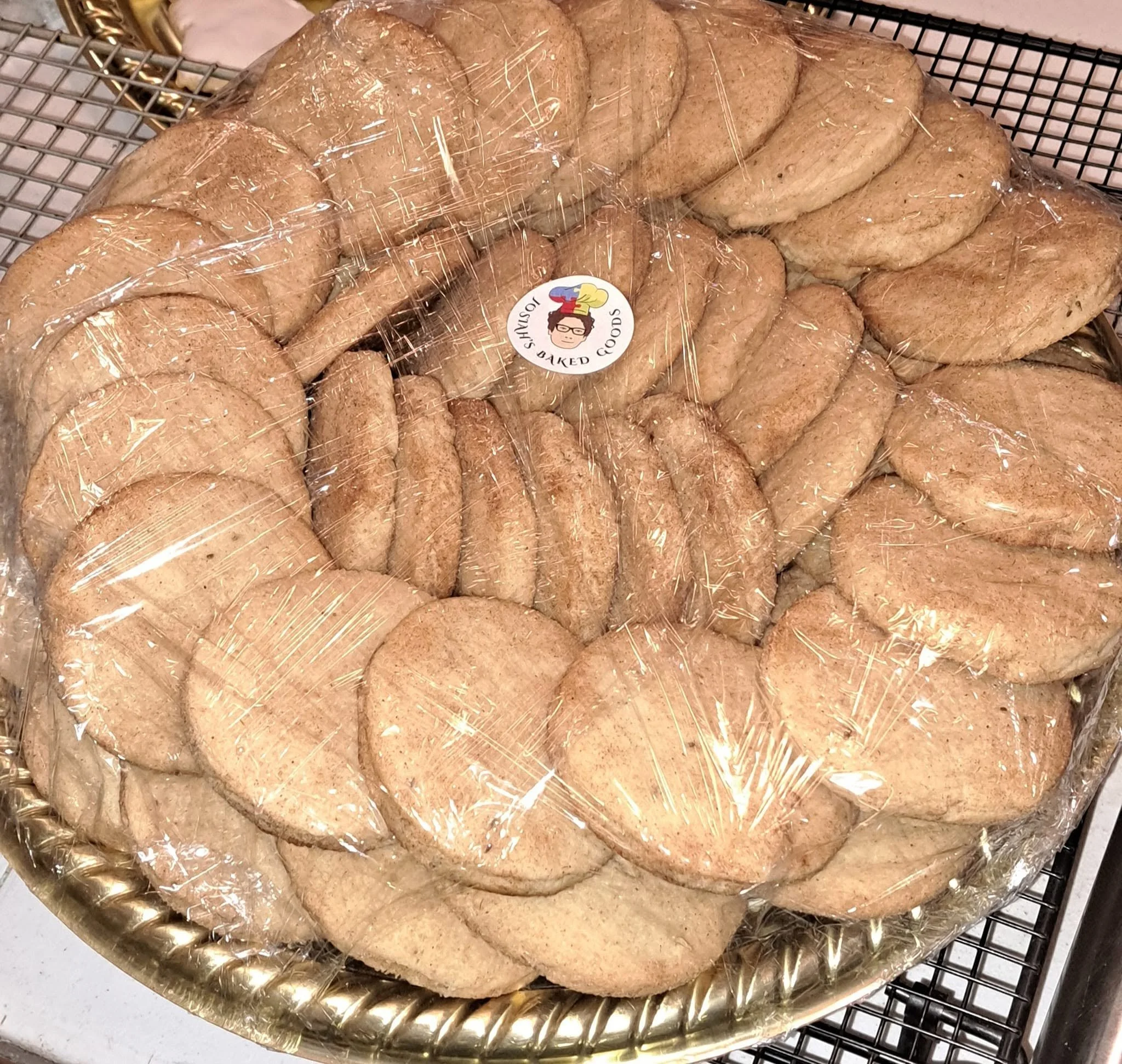 A large batch of baked cookies wrapped in plastic, placed on a gold-colored tray on a cooling rack.