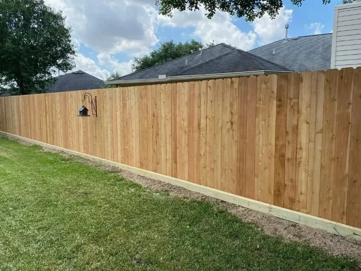 Side view of a newly installed wooden privacy fence with a grassy yard and a house roof in the background under a partly cloudy sky.