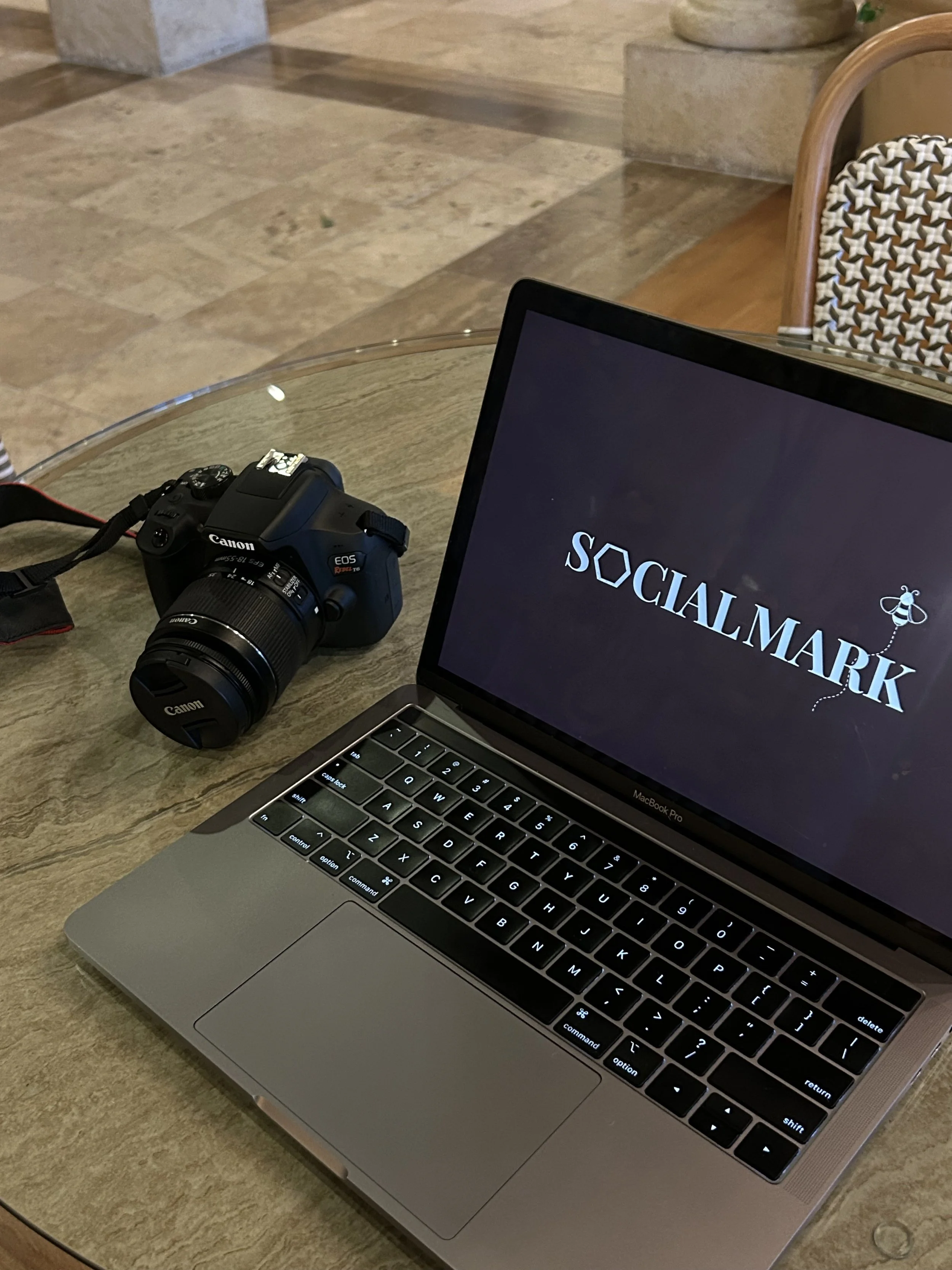 A MacBook Pro laptop on a wooden table with a partially visible screen displaying the word 'SOCIALMARK'. To the left of the laptop, there is a Canon DSLR camera with a lens and strap. In the background, there is a chair with a woven backrest and a patterned cushion, and floor steps made of stone or concrete.